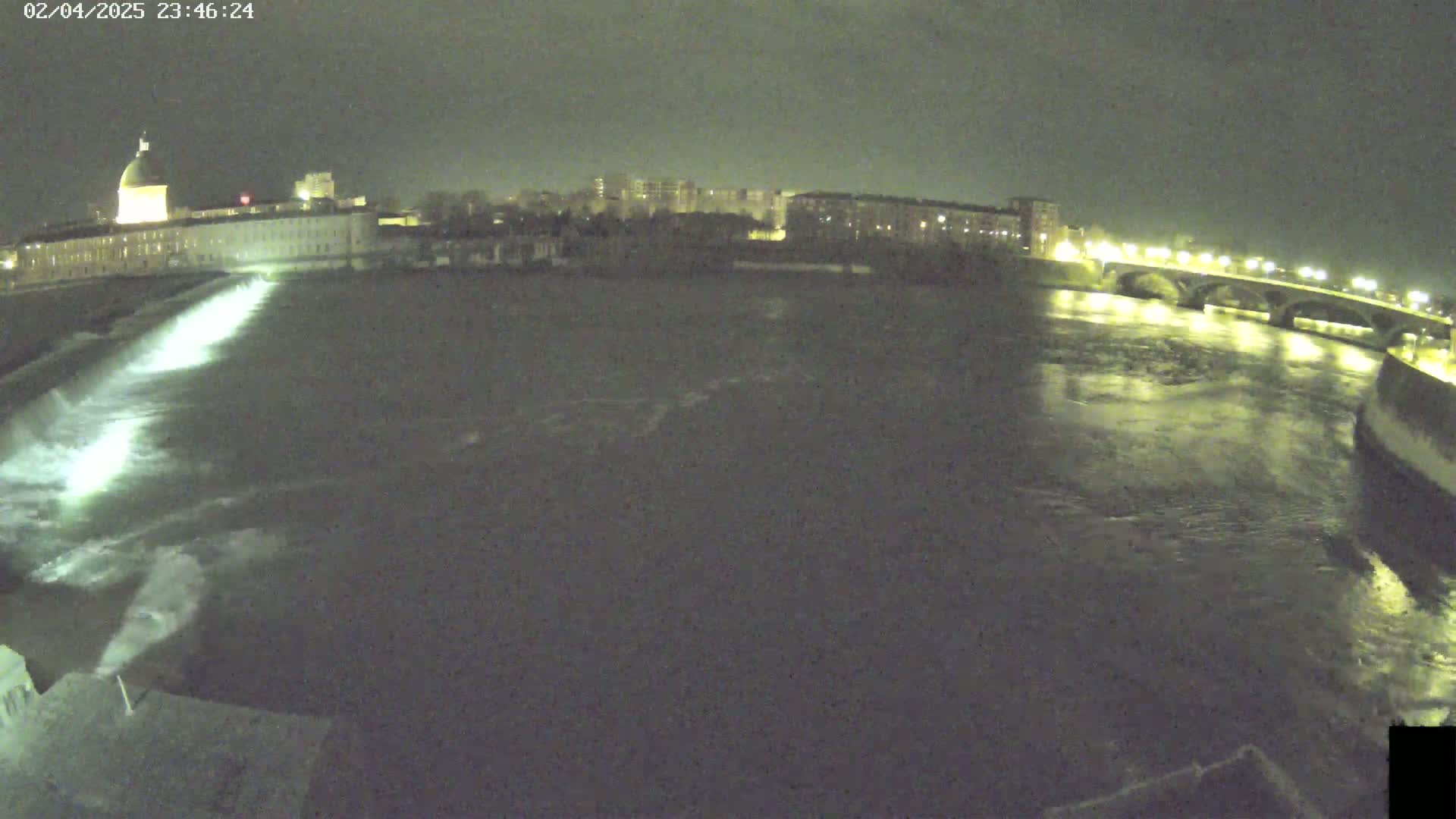 A nighttime view of a river flowing past a city with a building featuring a dome, a bridge illuminated with lights, and water flowing rapidly near a dam.