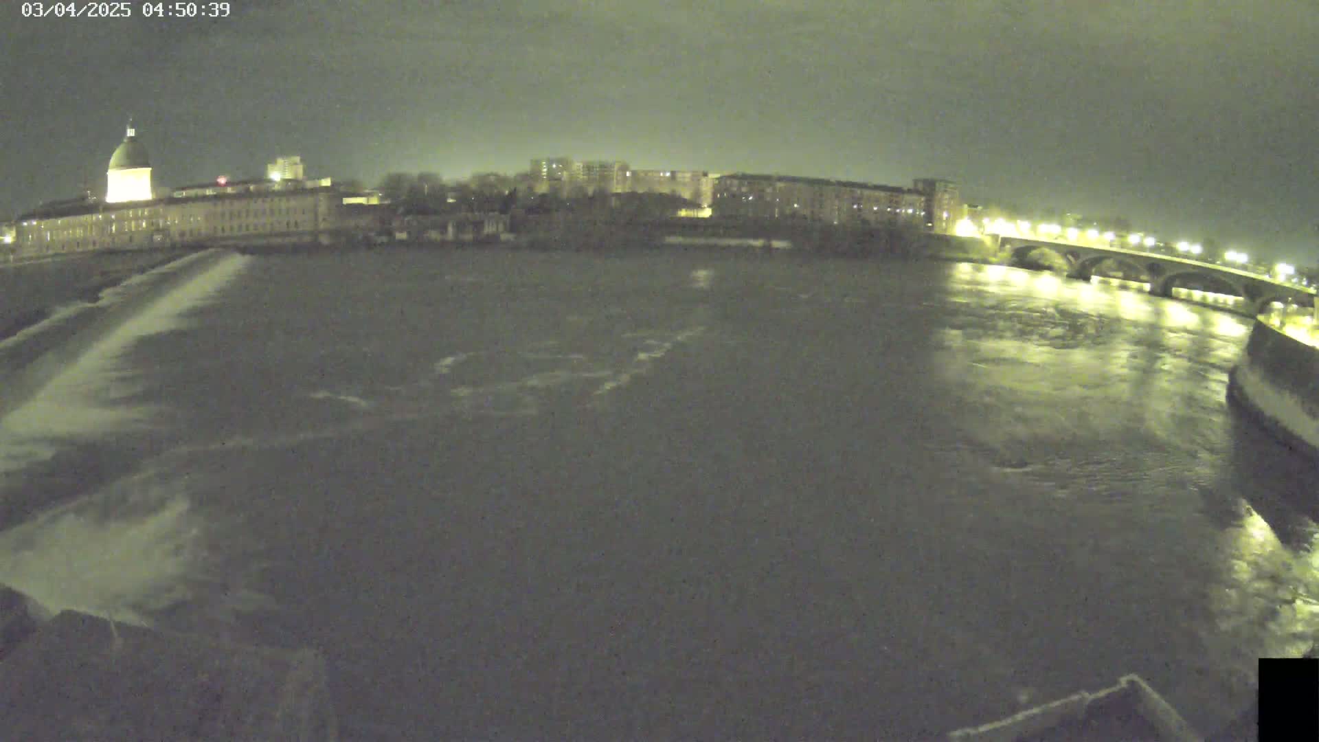 A nighttime view of a river flowing past a city, with a bridge visible in the distance under a dark sky.