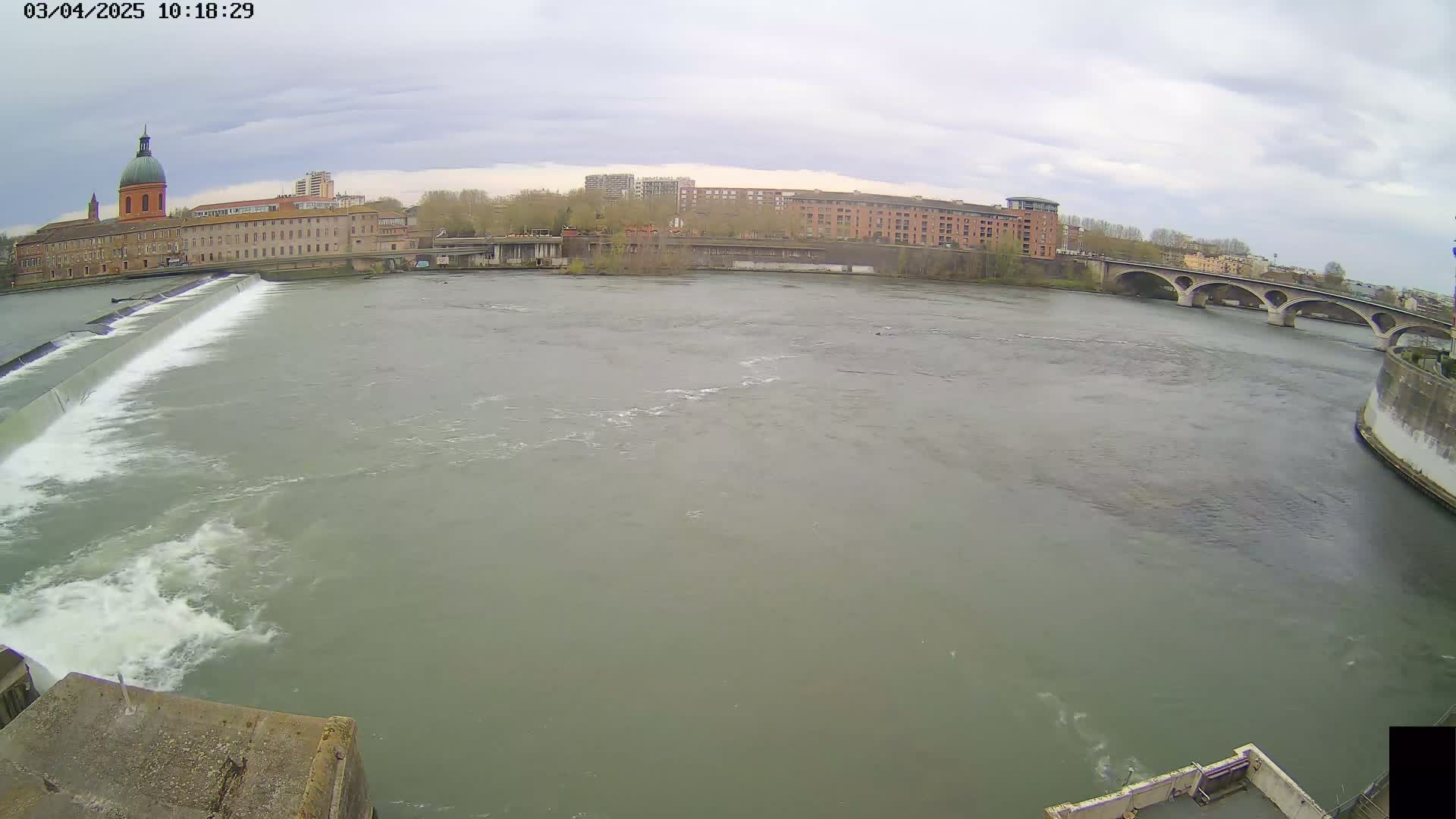 A wide shot of a river flowing under an overcast sky past a dam and buildings, including a domed building on the left bank and a bridge on the right.