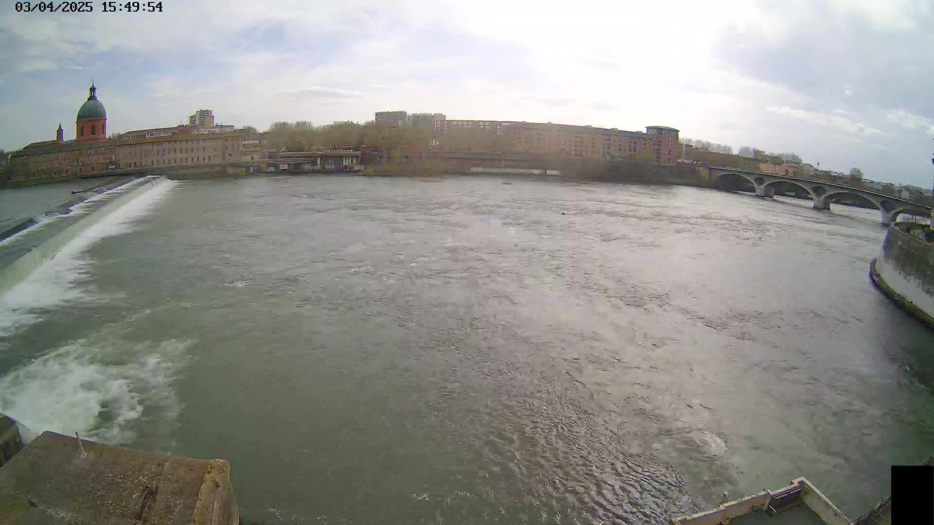 A wide shot shows a river flowing past a dam and under a bridge, with buildings and a dome-topped structure visible on the far bank under a partly cloudy sky.