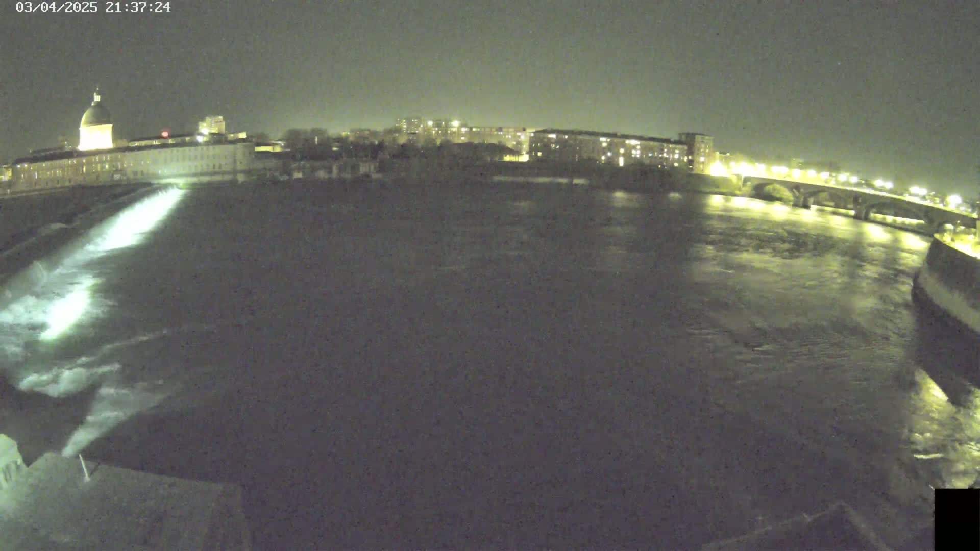 A nighttime view of a river flowing past a city, with a dam releasing water, under a dark sky.