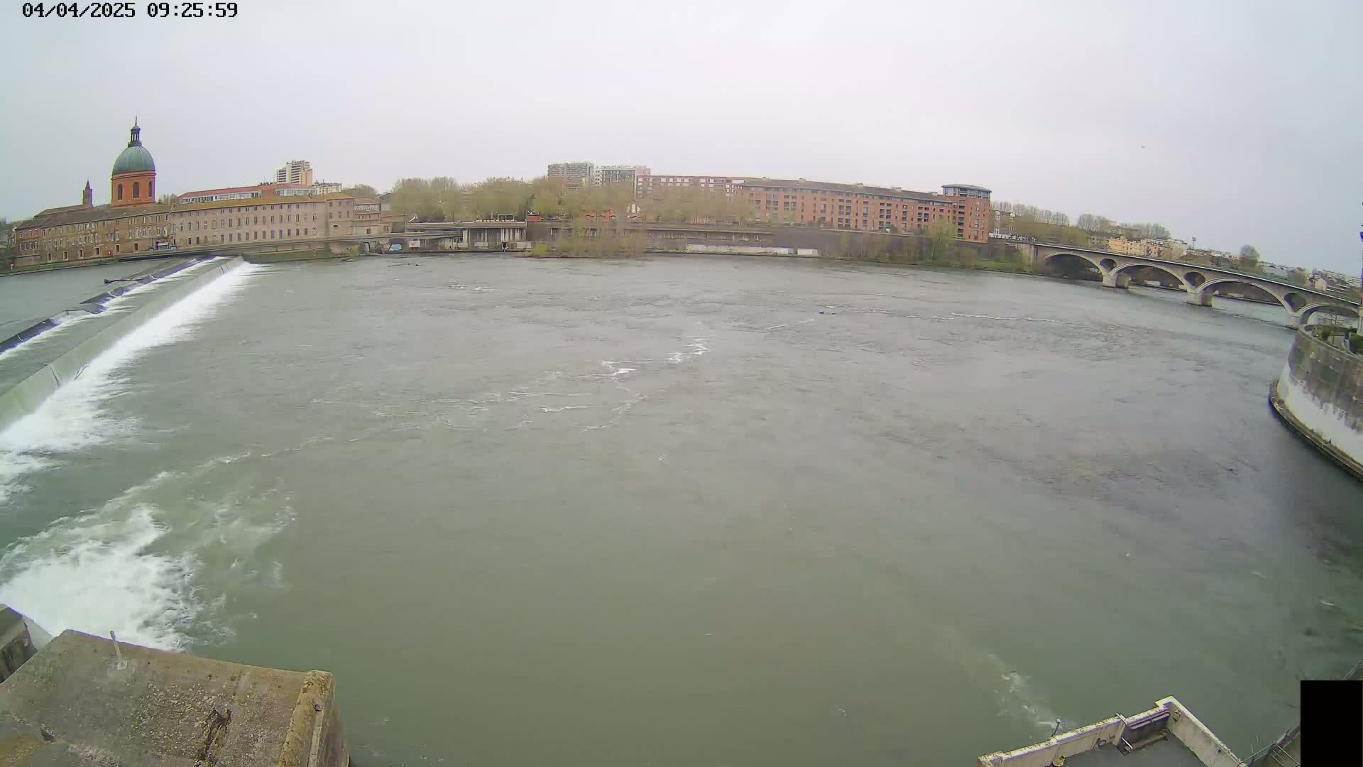 A wide shot of a river flowing past a city on an overcast day, featuring a dam, buildings, and an arched bridge.