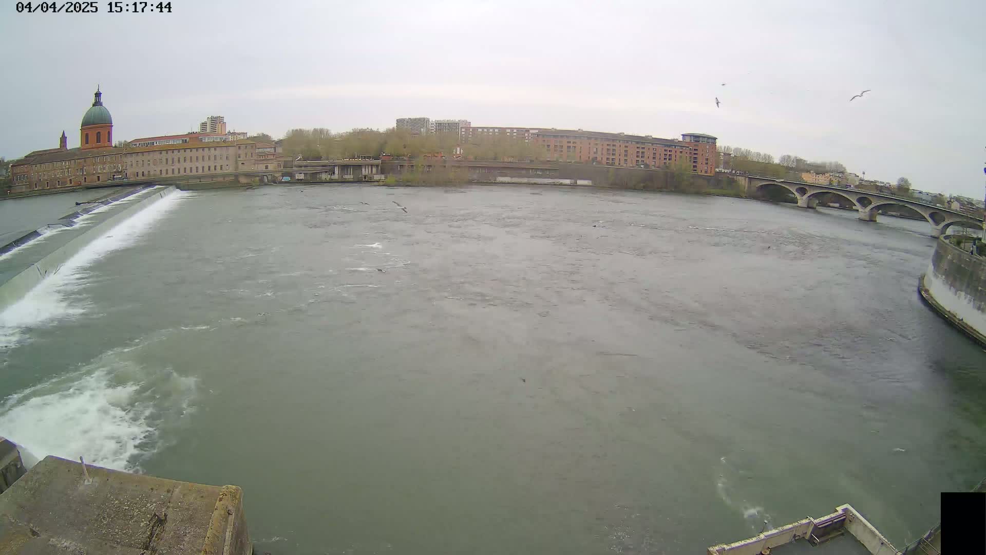 A wide shot of a river flowing past a dam and under a bridge, with buildings and a domed structure visible on the far bank under an overcast sky.