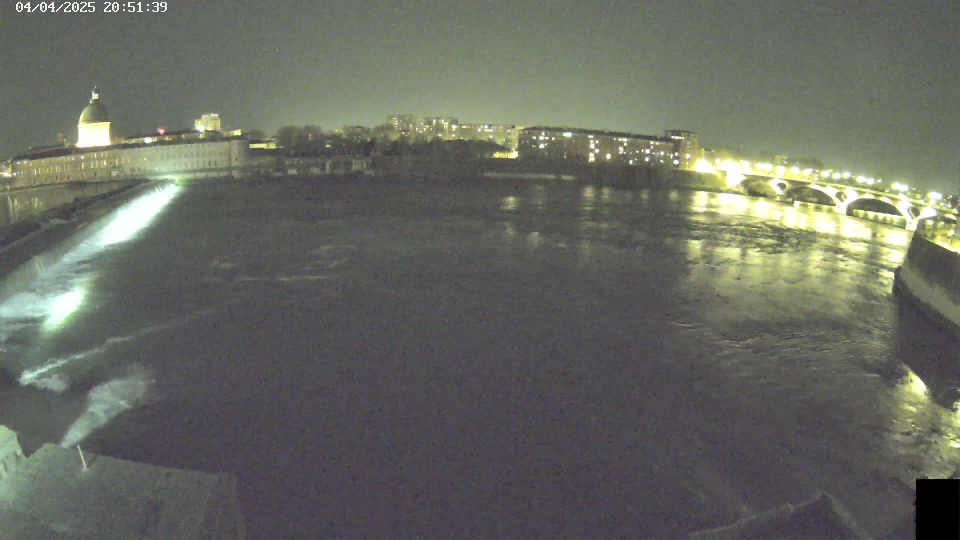 A nighttime view of a river flowing past a city, with a dam releasing water and a bridge illuminated in the distance.