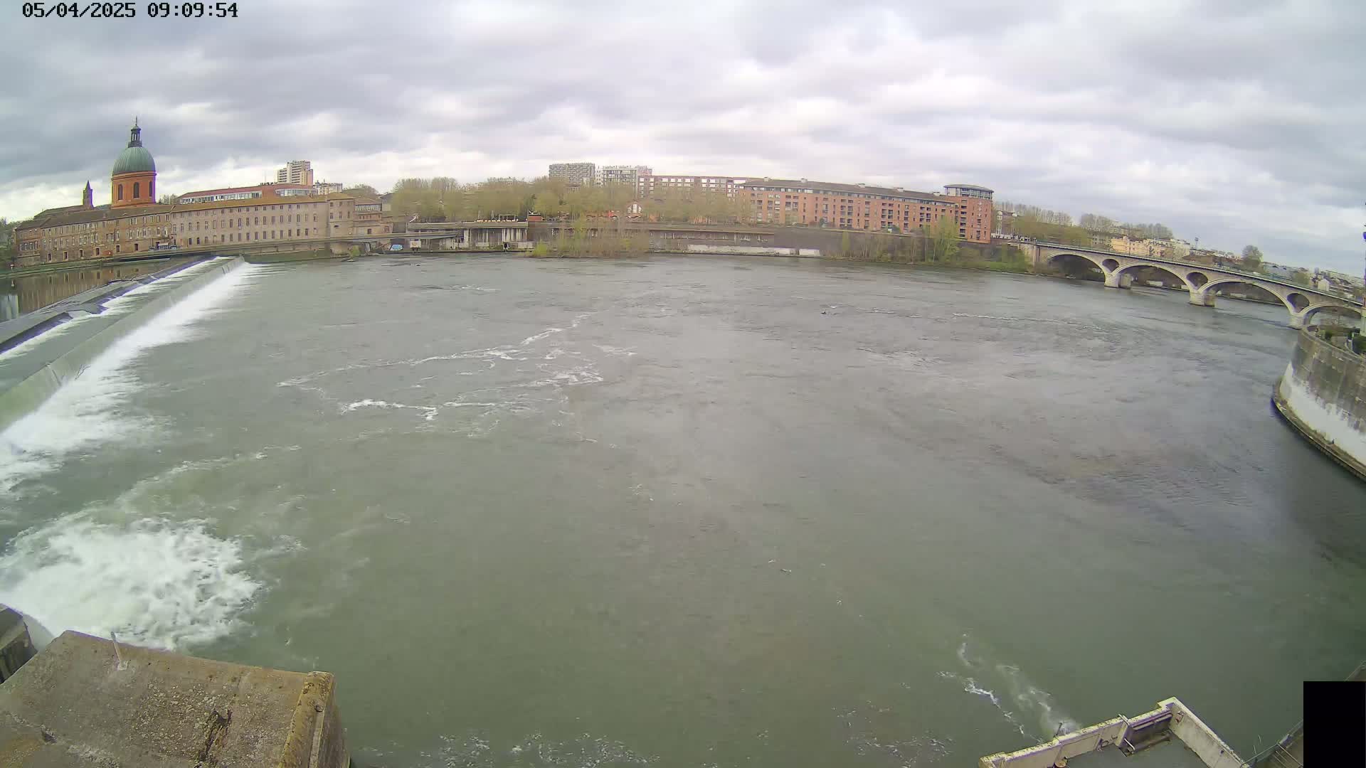 A wide shot of a river flowing past a dam and under a bridge, with overcast skies and buildings lining the riverbank.