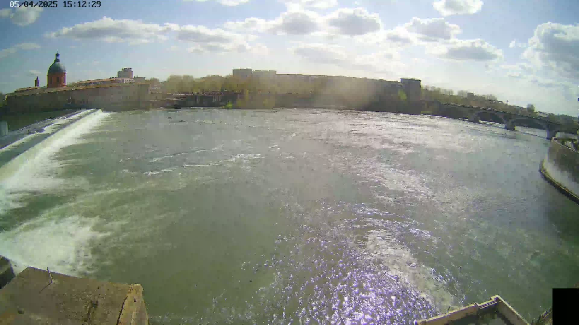 A river flows past a city under a partly sunny sky, with a dam and a bridge visible in the distance.