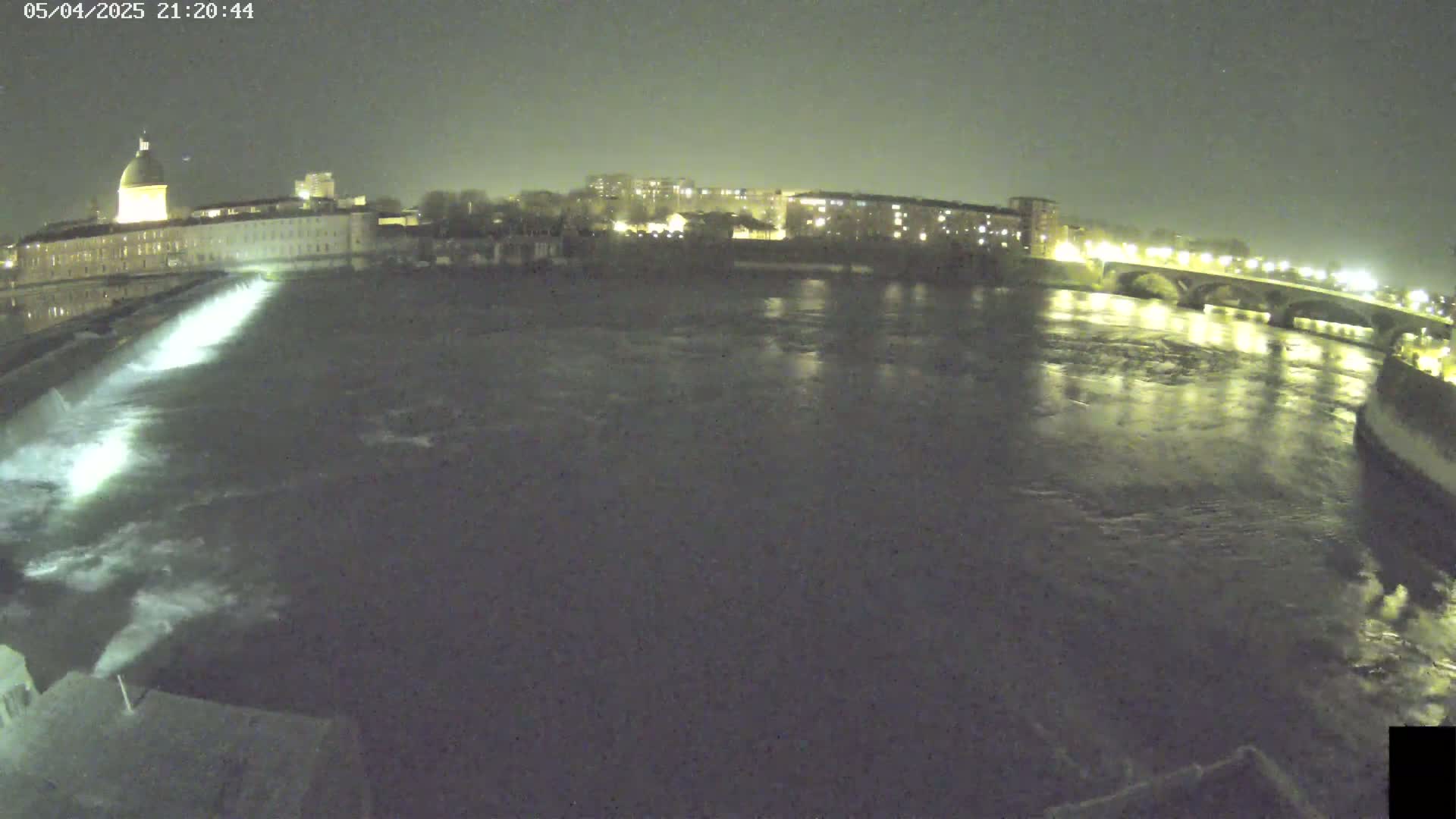 A nighttime view of a river flowing past a city, with a dam visible in the foreground and a bridge in the distance under a dark sky.