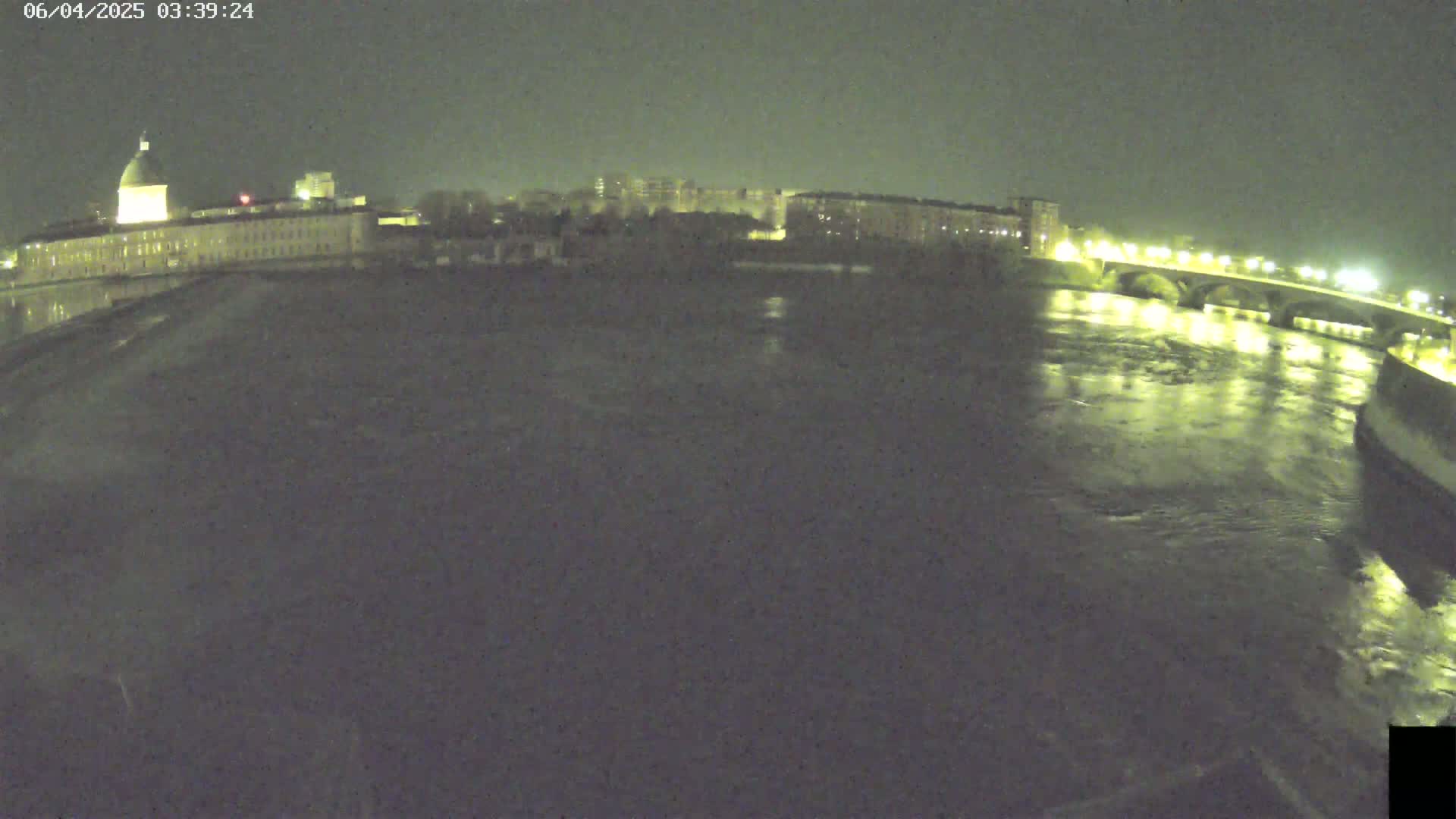 A nighttime view of a river flowing past a city with buildings and a bridge illuminated by lights.
