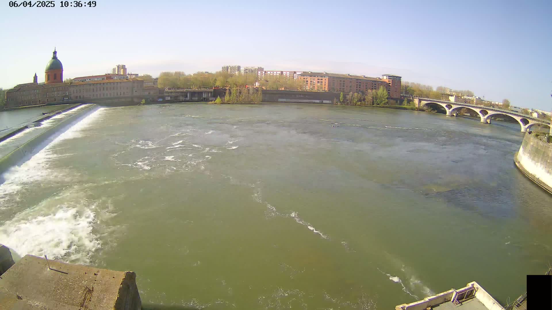 A wide shot of a river flowing under a bridge on a sunny day, with a dam and buildings visible in the background.