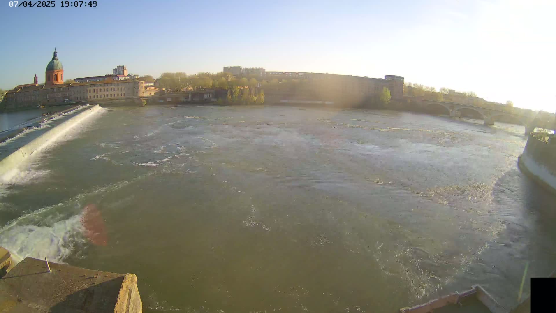 A wide, brown river flows past a stone dam and a city with buildings, including a domed structure, under a clear, sunny sky.