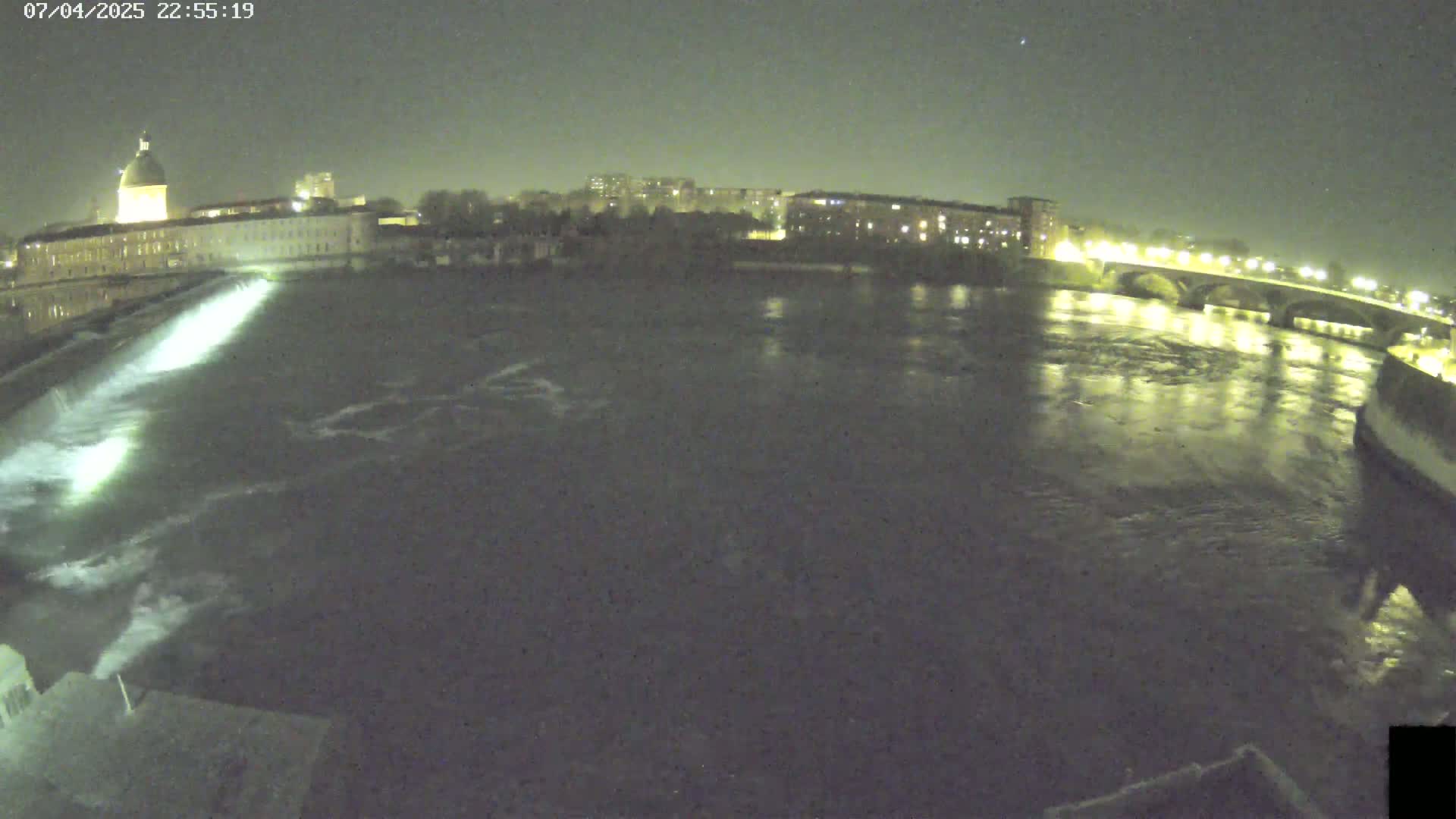 A nighttime view of a river flowing past a city, with a dam and bridge visible under a clear sky.