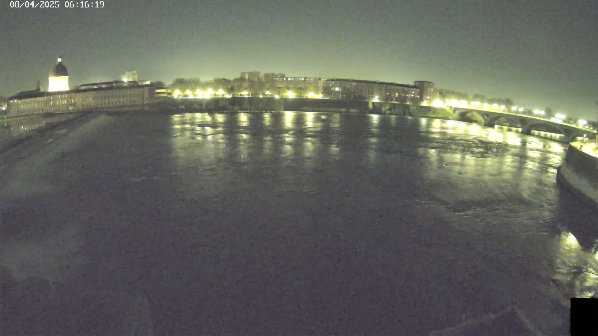 A wide nighttime shot shows a dark river reflecting the lights of buildings and a bridge on the far shore.