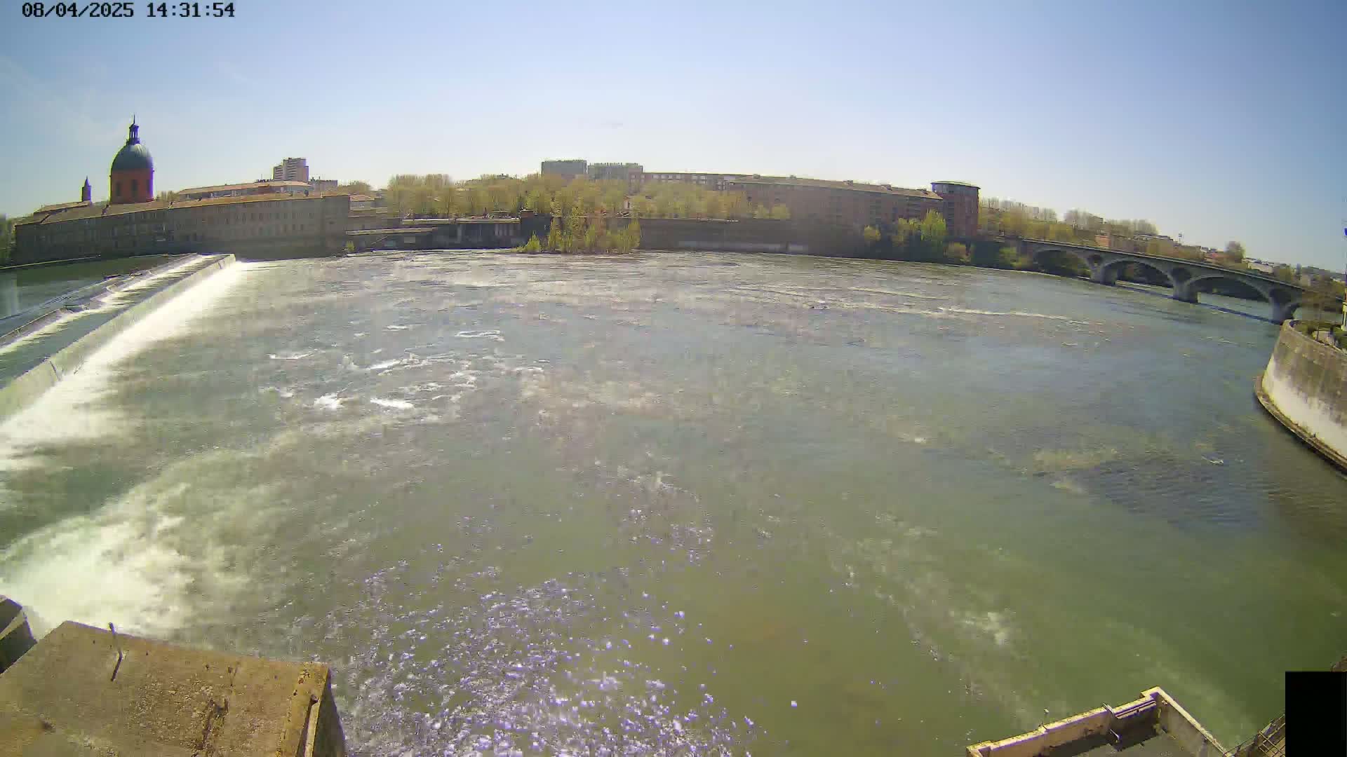 A wide shot of a river flowing under a bridge on a sunny day, with a dam and buildings visible in the background.