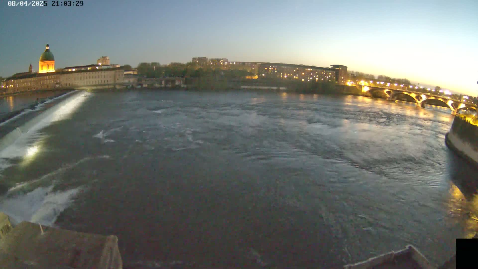 A wide shot at dusk shows a river flowing past a dam and under a bridge, with city buildings and lights visible on the far bank.