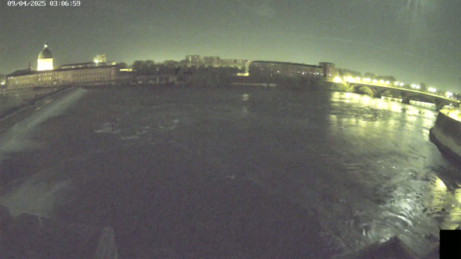 A nighttime view of a river flowing past a city with buildings, including a large domed structure, and a bridge, under a dark sky.