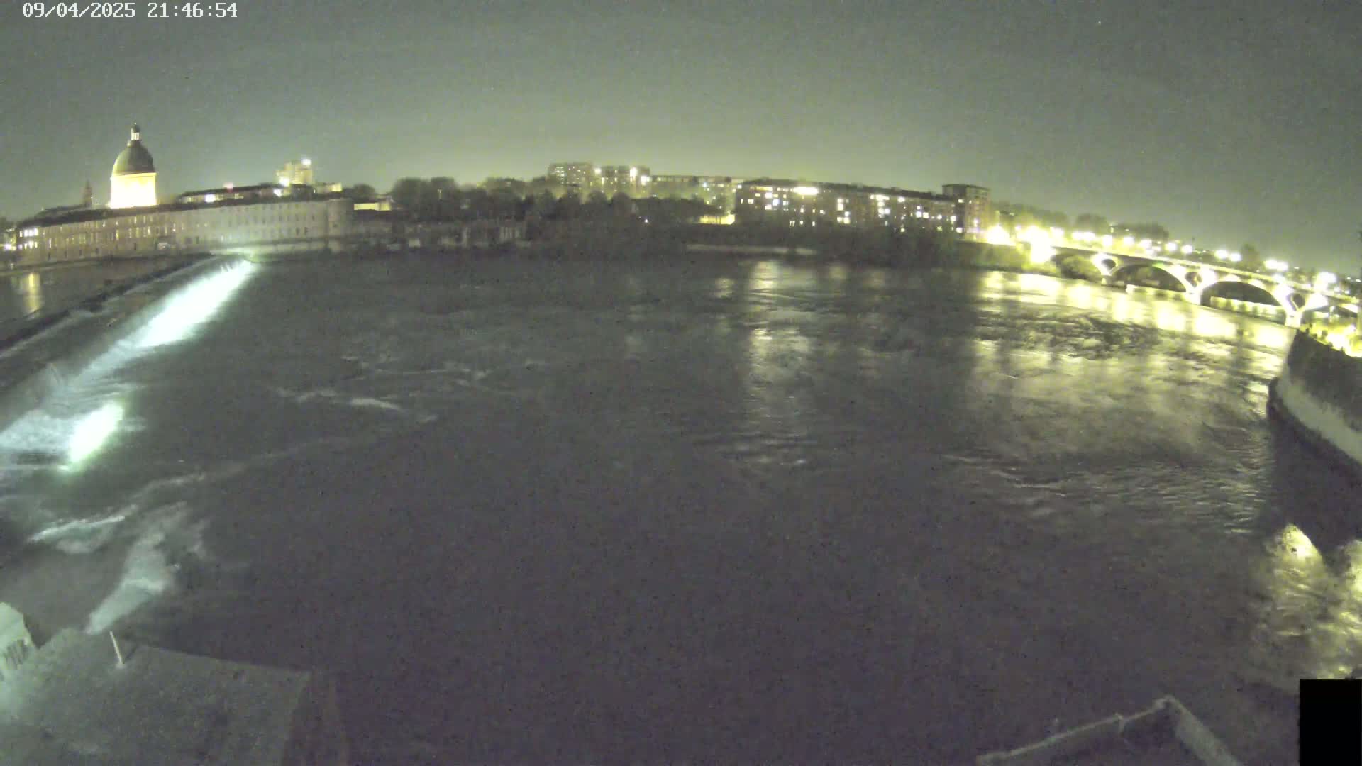 A nighttime view of a river flowing past a city, with a dam and bridge visible under a dark sky.