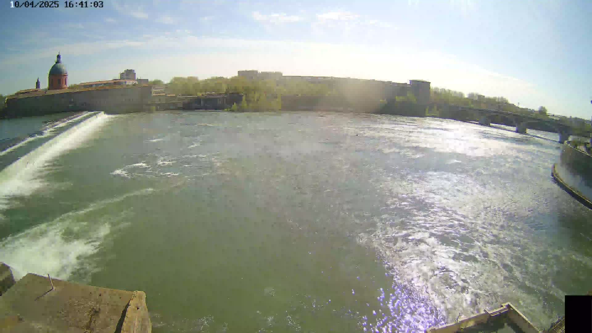 A wide shot of a river flowing past a dam and buildings on a sunny day.