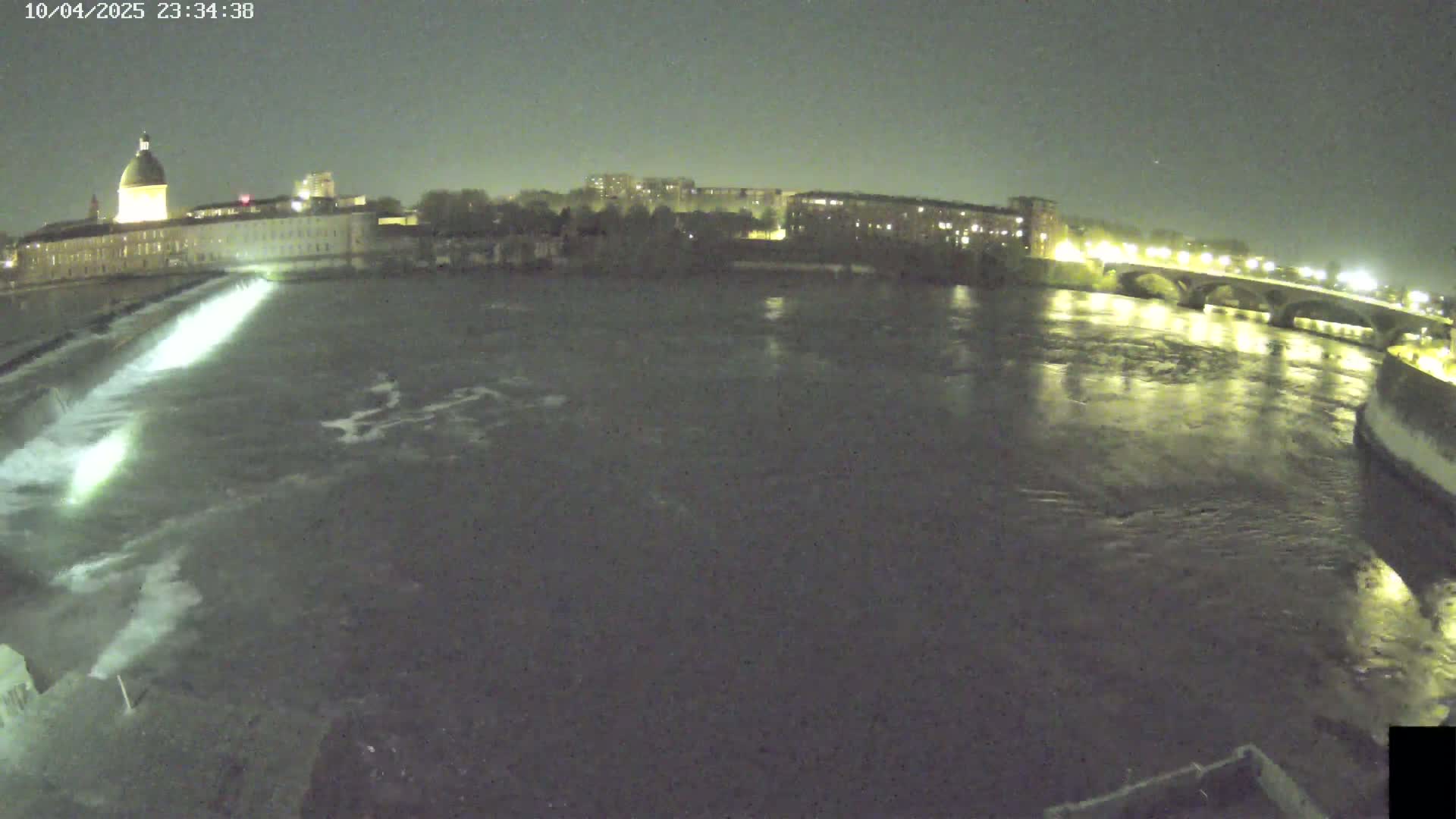 A nighttime view of a river flowing past a city, with a dam visible in the foreground and a bridge in the distance under a dark sky.