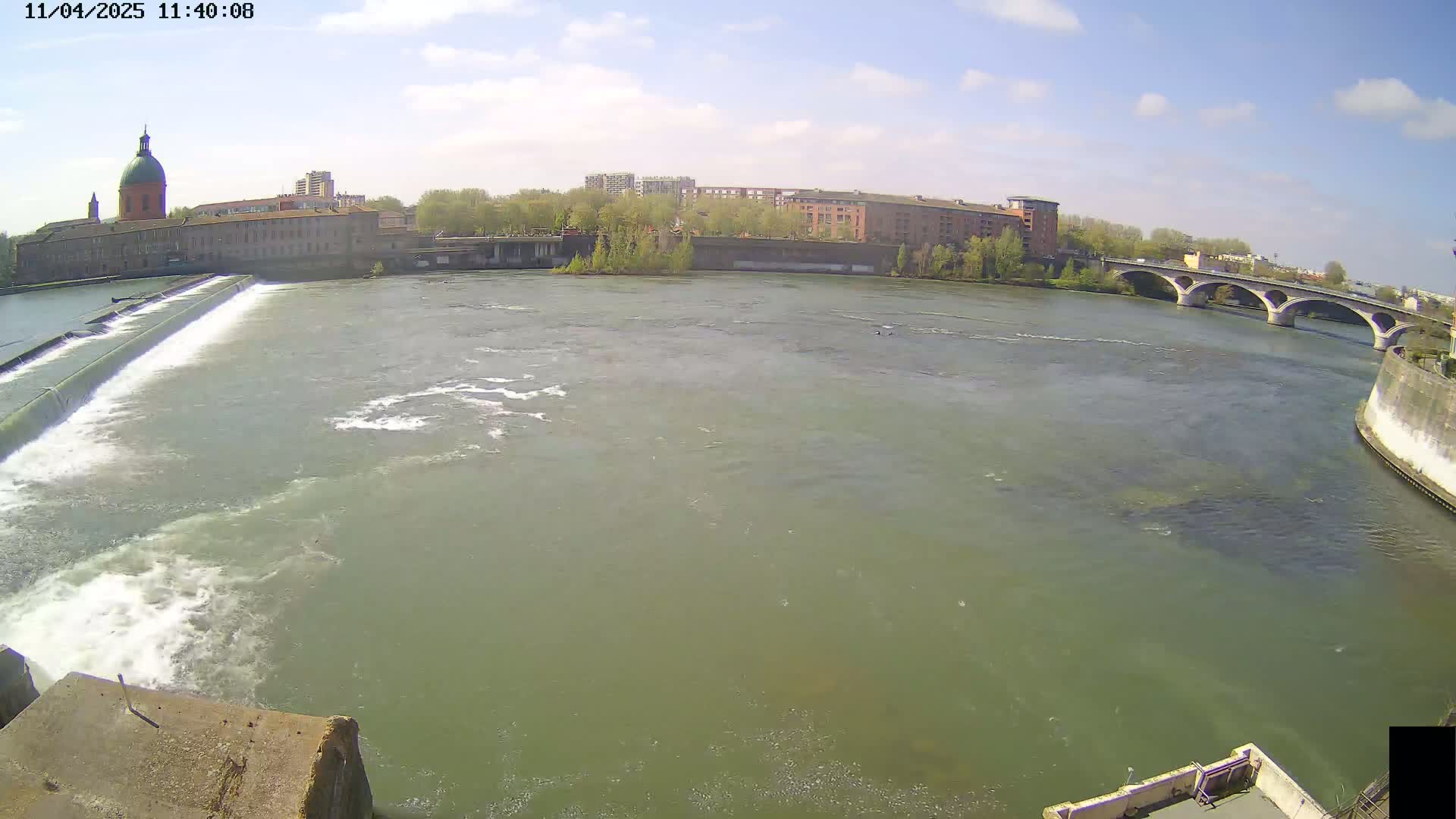 A wide shot of a river flowing under a bridge on a partly sunny day, with a dam and buildings visible in the background.