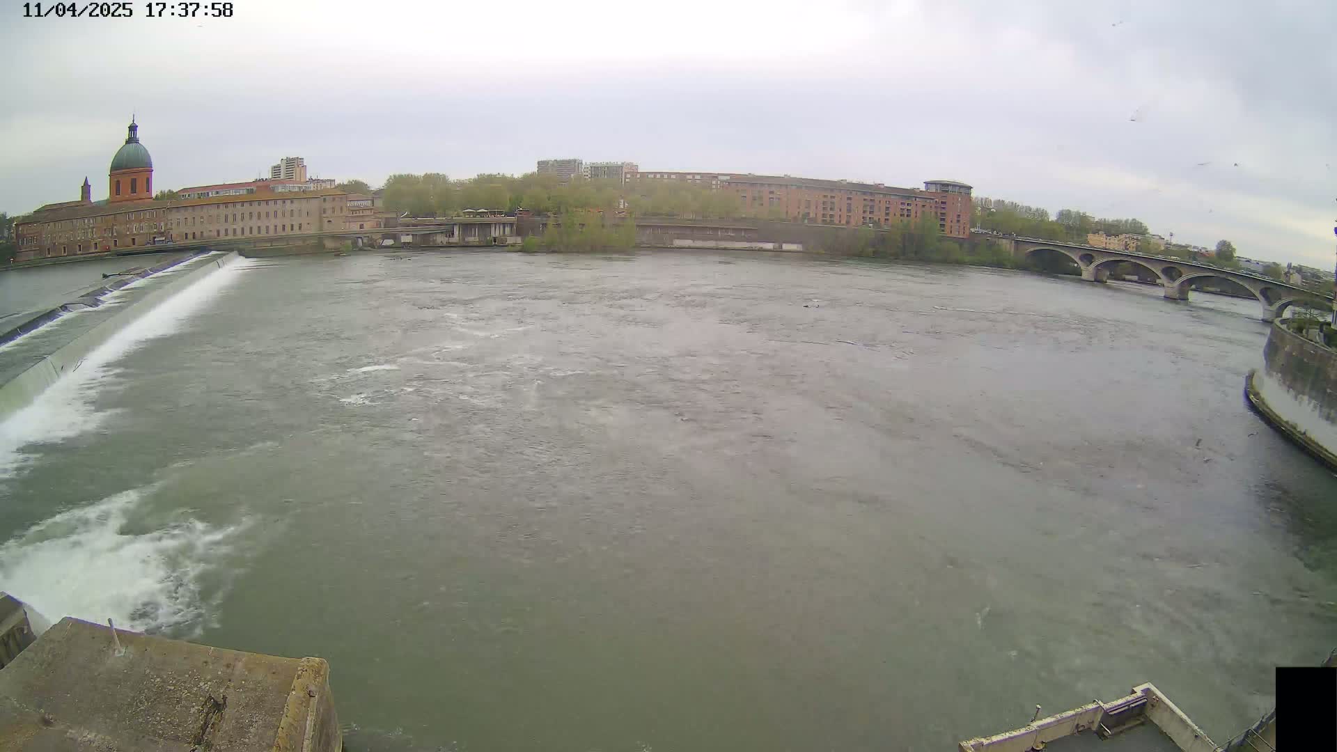 A wide shot of a river flowing past a city under an overcast sky, with a dam and bridge visible in the foreground and background, respectively.