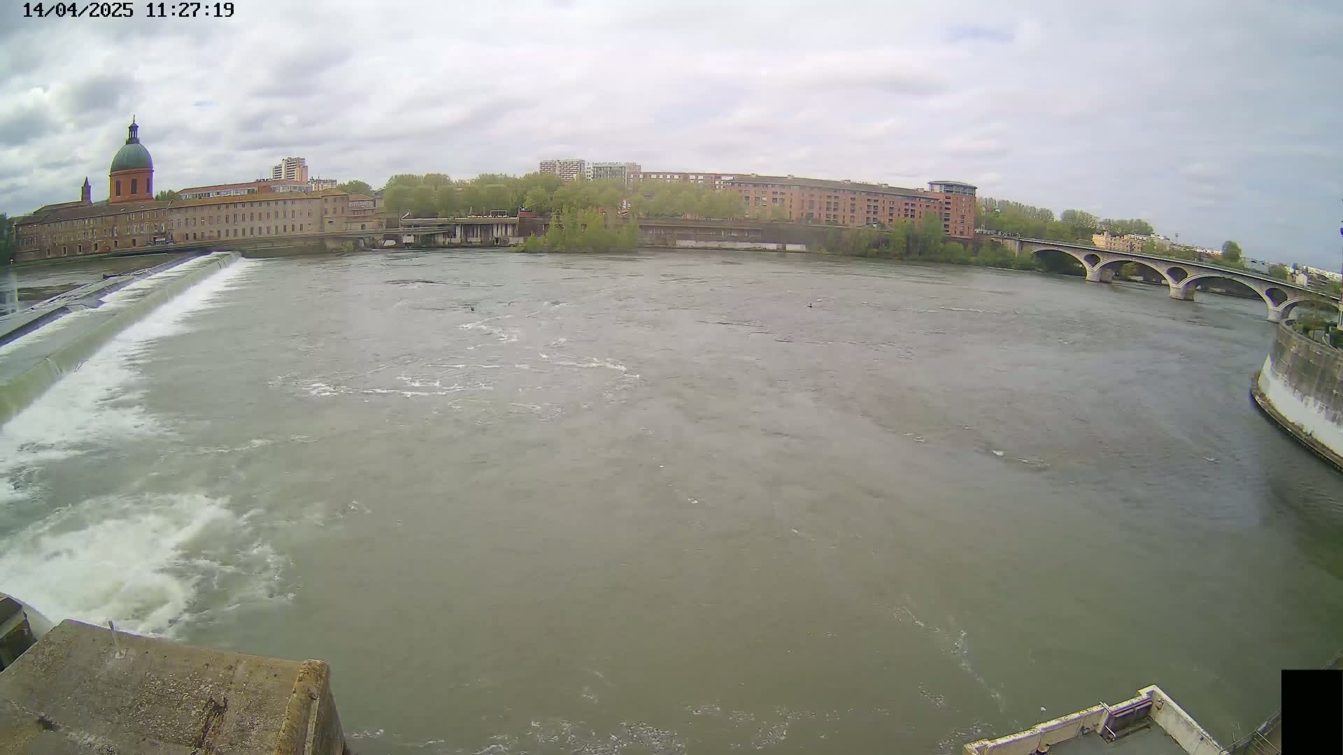 A wide shot of a river flowing past a dam and under a bridge, with buildings and trees lining the banks under a cloudy sky.