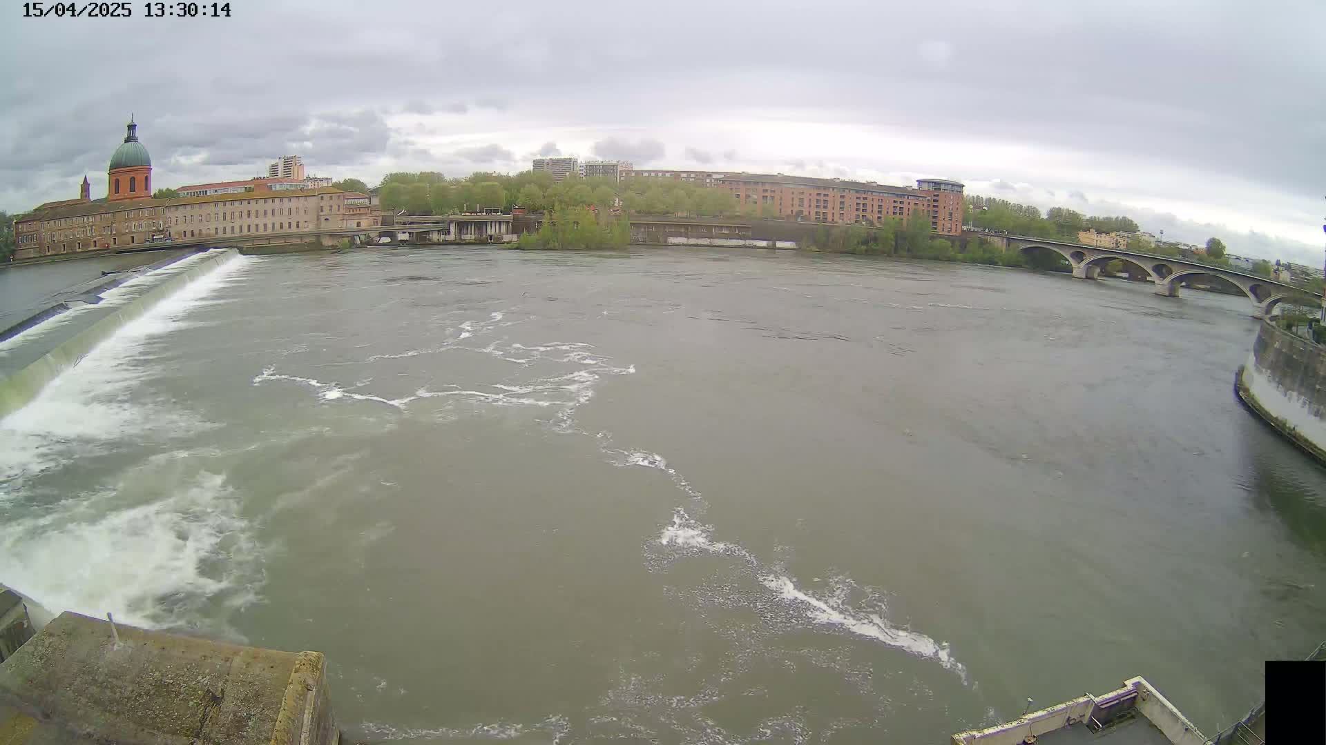 A wide shot shows a river flowing under an overcast sky past a dam and buildings, including a domed building and a bridge.