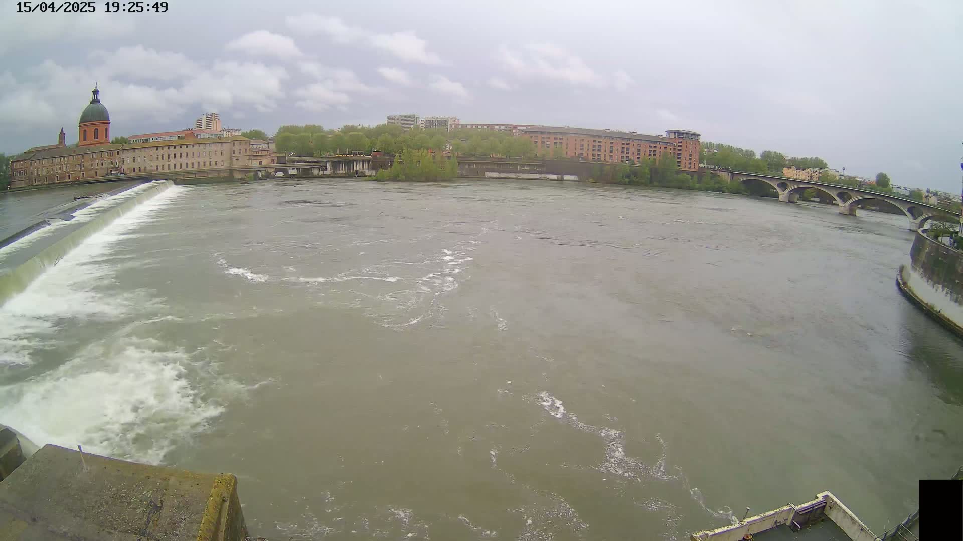 A wide river flows under an overcast sky past a dam, buildings, and a bridge.