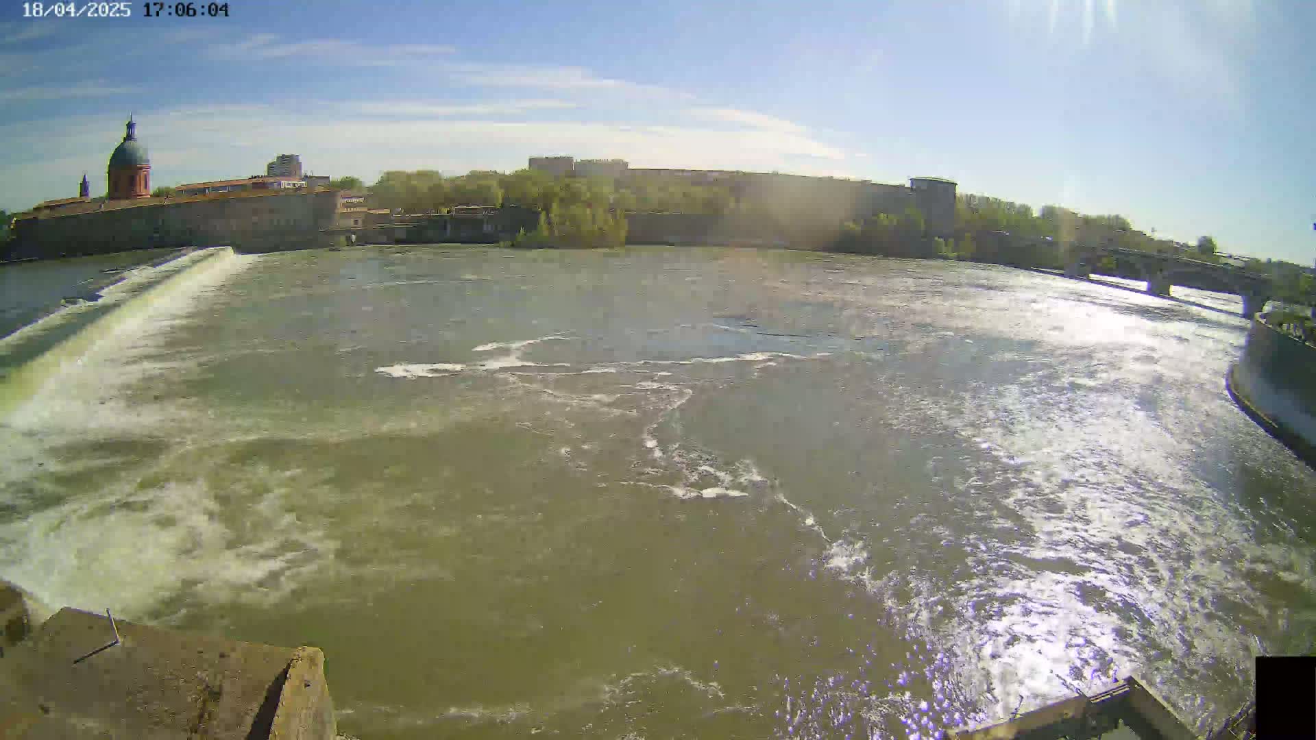 A wide shot of a river flowing past a dam and city buildings under a sunny sky.