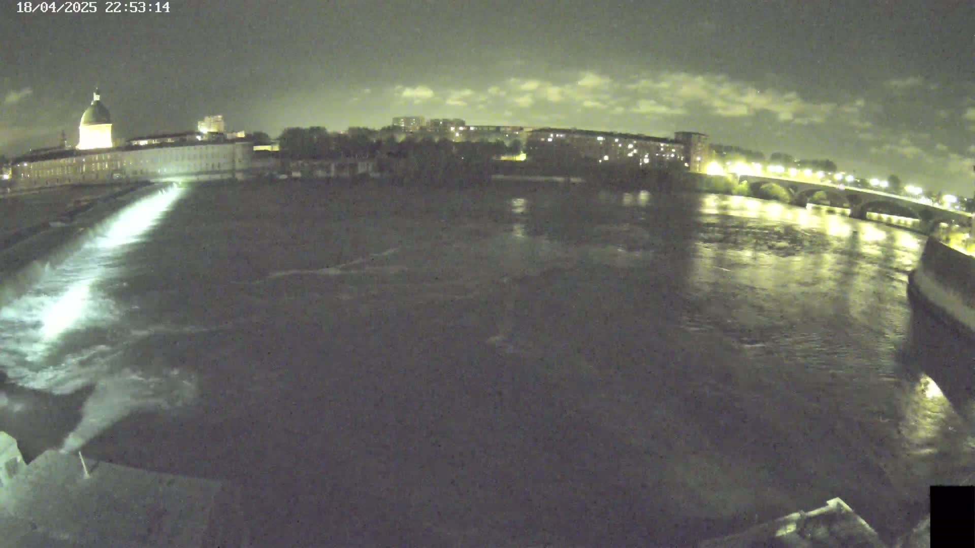 A nighttime view of a river flowing past a city with buildings, a bridge, and a dam releasing water.