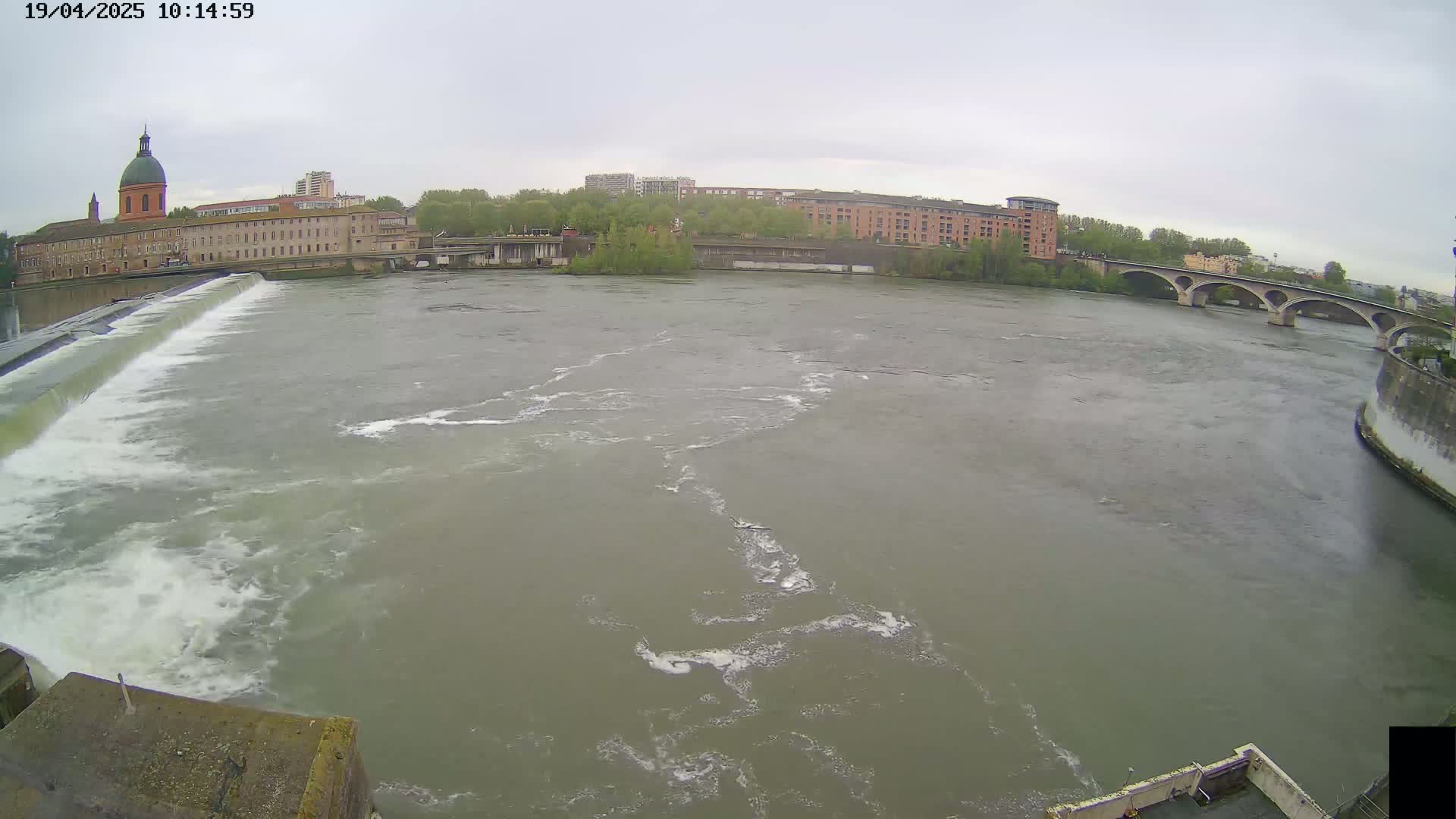 A wide shot of a river flowing past a dam and buildings under an overcast sky.