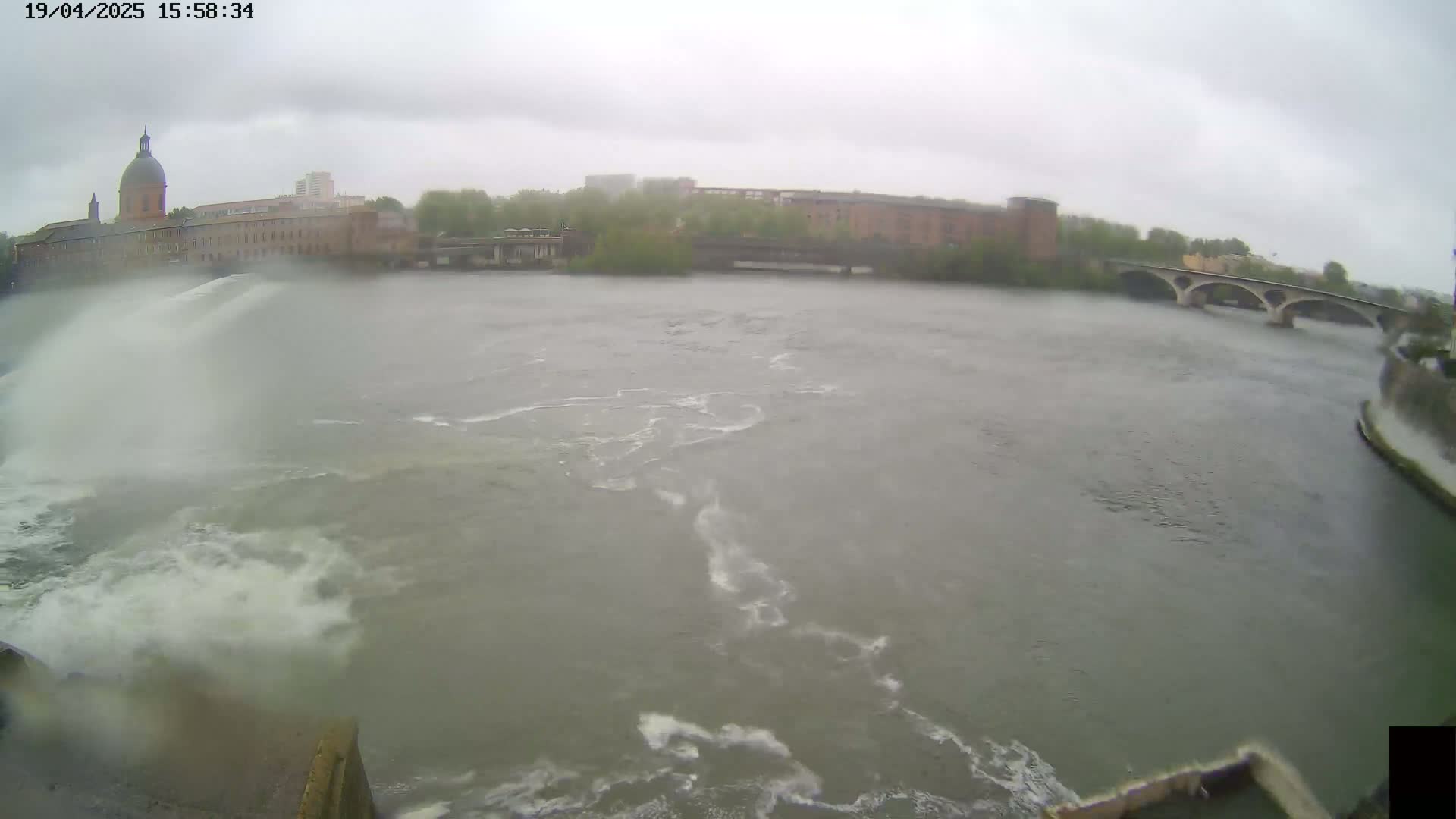 A wide shot of a river on an overcast day shows churning water near a dam and a bridge in the distance, with buildings and trees along the riverbank.