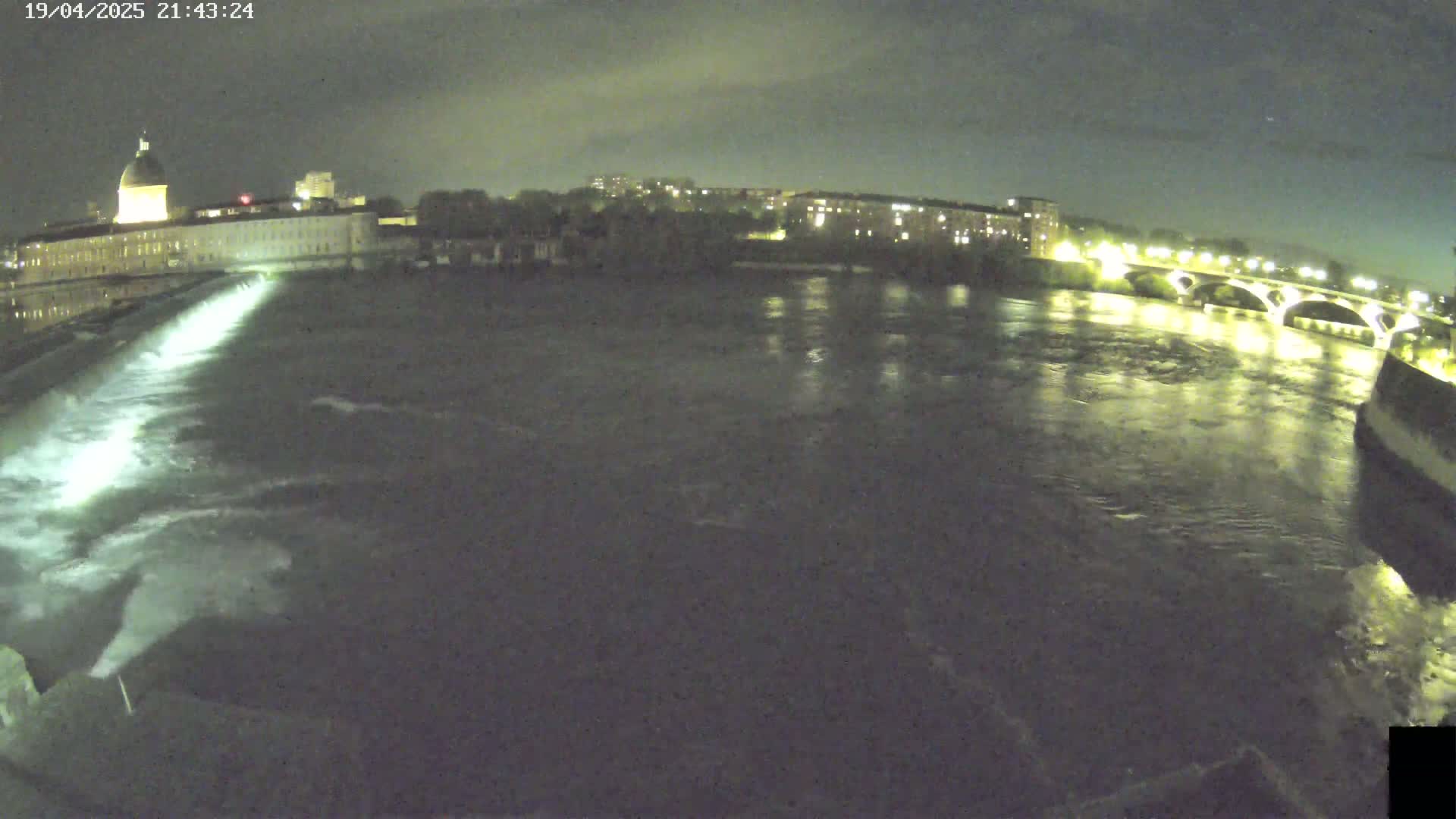 A nighttime view of a river flowing past a city, with a dam and bridge illuminated under a partly cloudy sky.