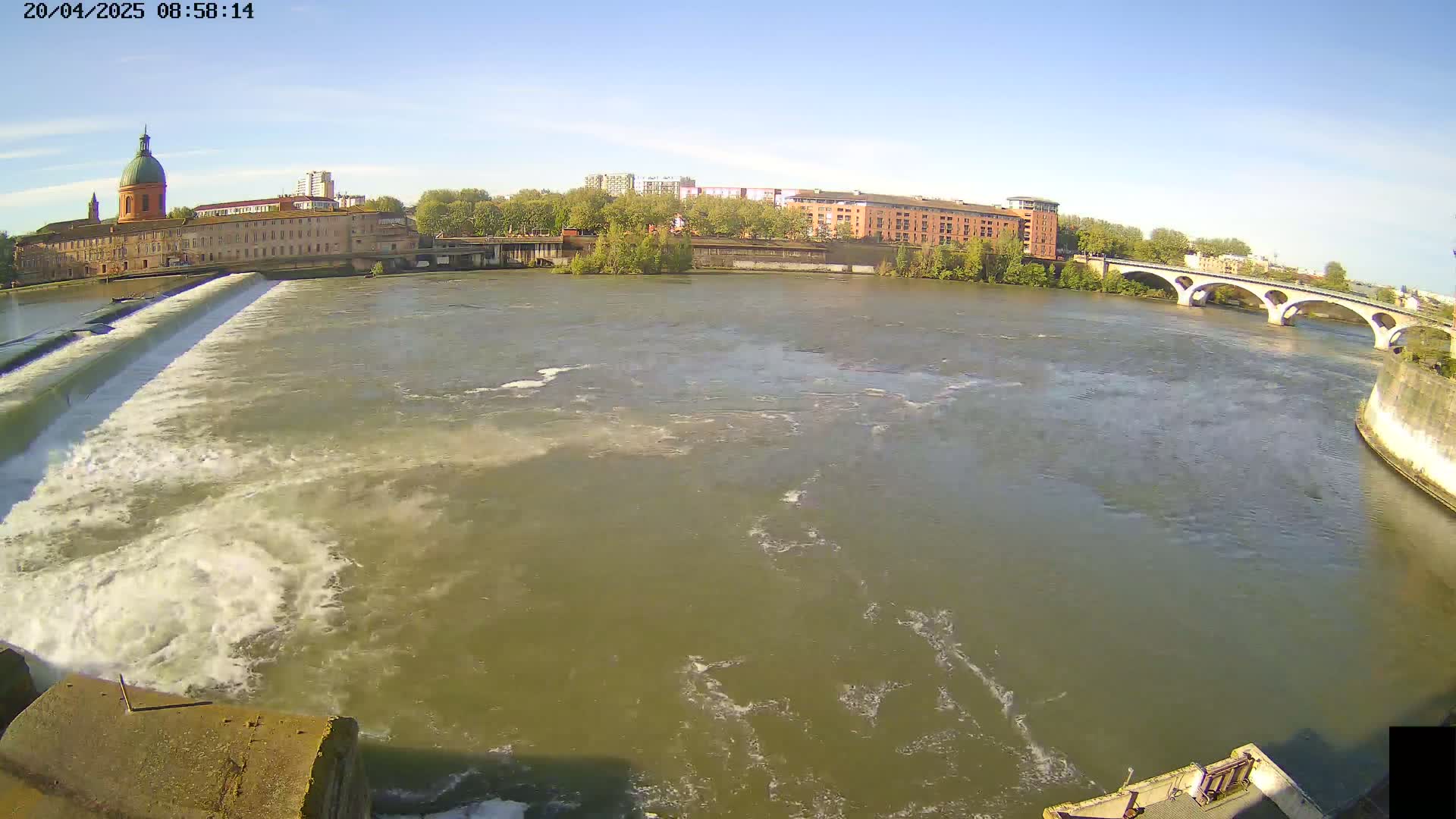 A wide shot of a river flowing under a bridge past buildings and a dam on a sunny day.