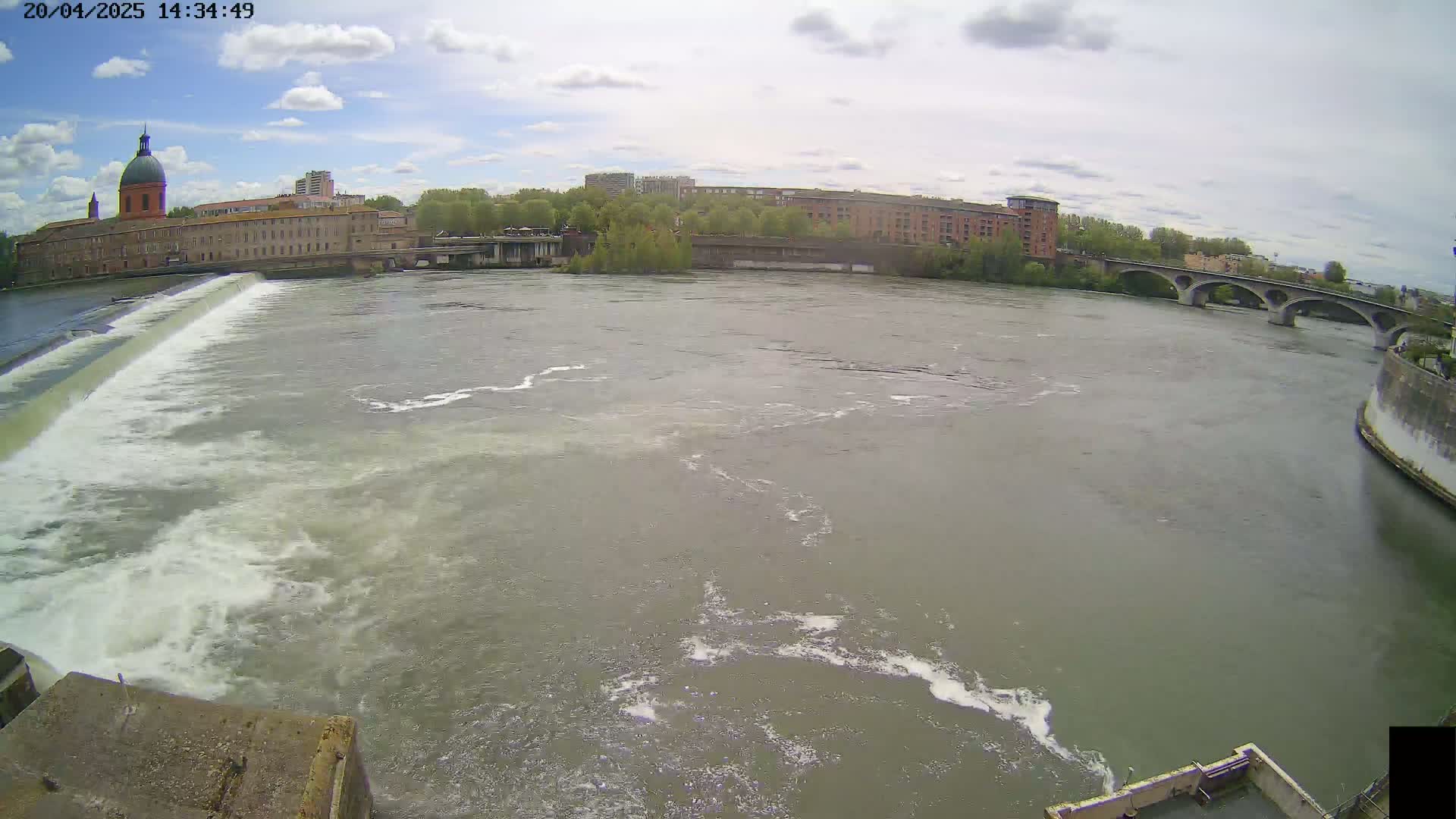 A wide shot of a river flowing under a bridge past buildings, with a dam creating whitewater on the left side under partly cloudy skies.