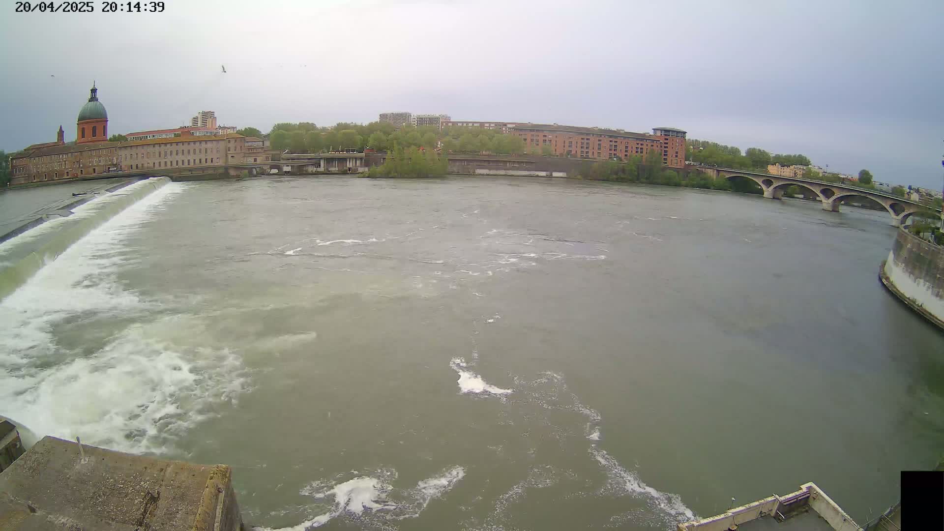 A wide shot of a river flowing past a dam and under a bridge on an overcast day, with buildings and trees visible on the shore.