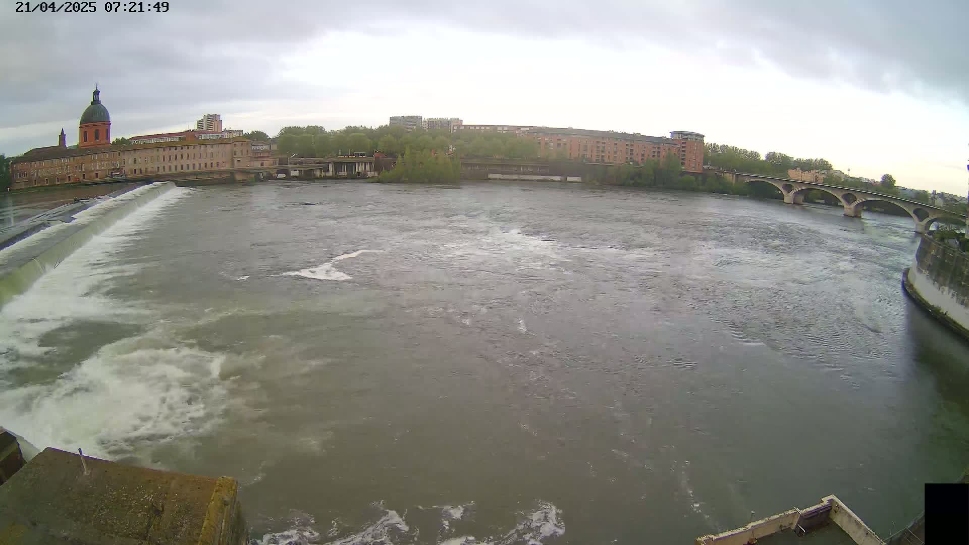 A wide shot of a river flowing past a dam and under a bridge on an overcast day, with buildings visible in the background.
