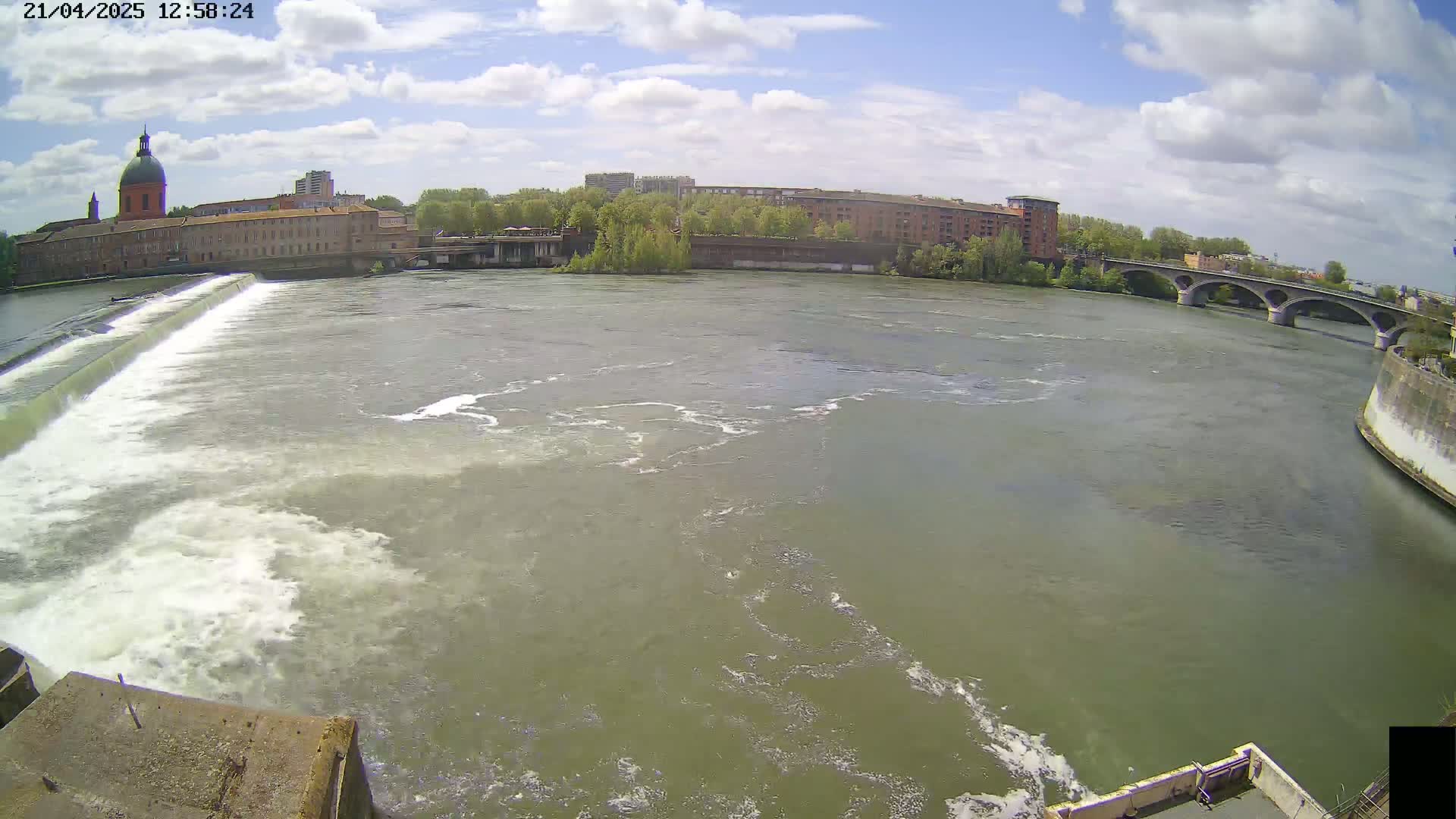 A wide shot of a river flowing under a bridge on a partly cloudy day, with a dam and buildings visible in the background.