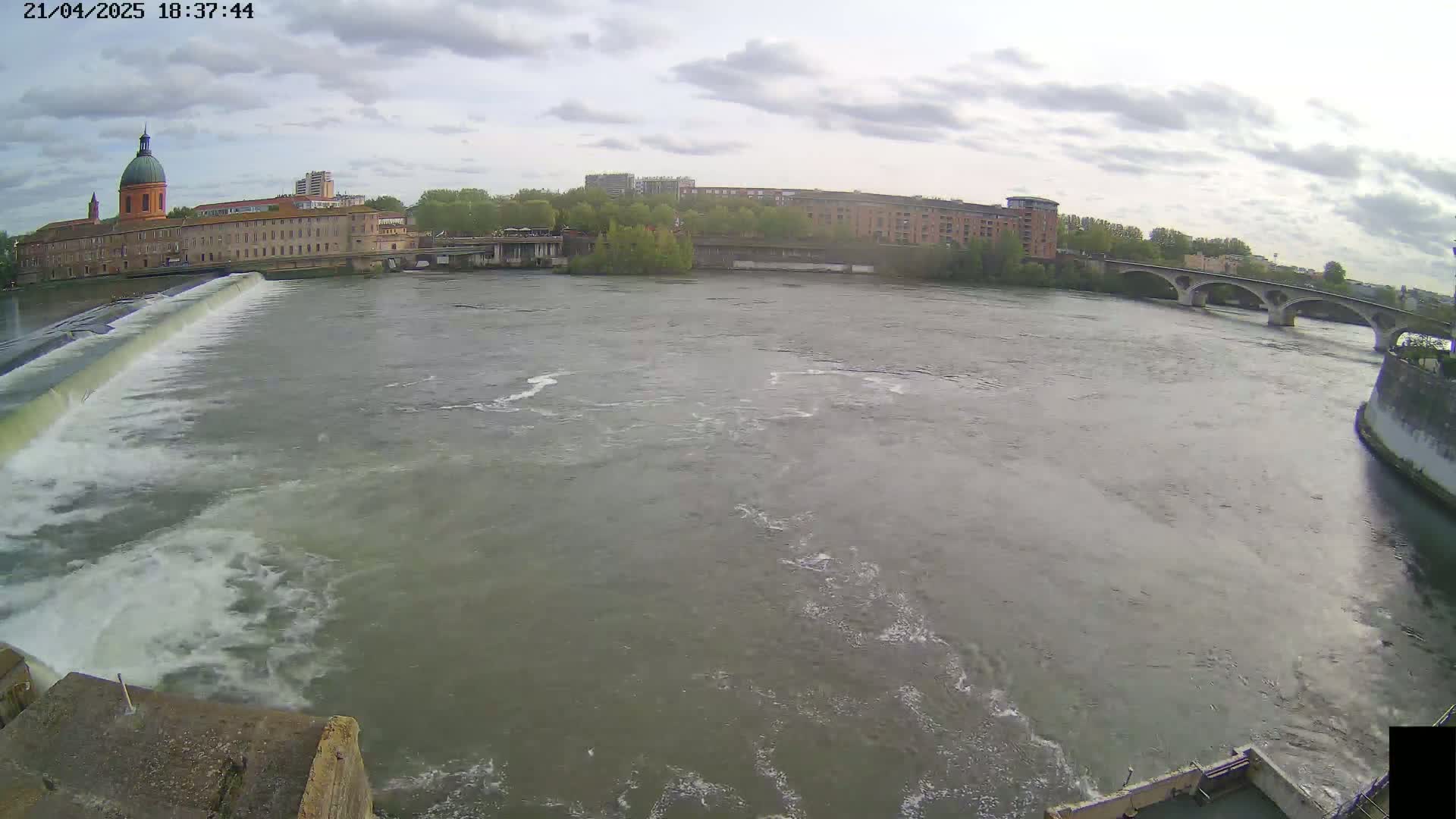 A wide shot of a river flowing past a dam and under a bridge, with buildings and trees visible on the far bank under a cloudy sky.