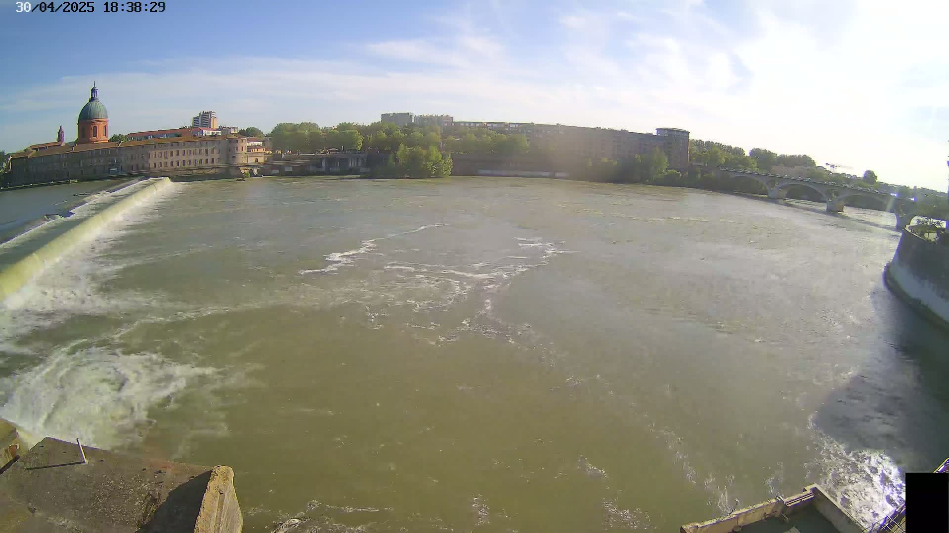 A wide shot shows a river flowing past a dam and under a bridge, with a large building and other structures visible on the far bank under a partly sunny sky.