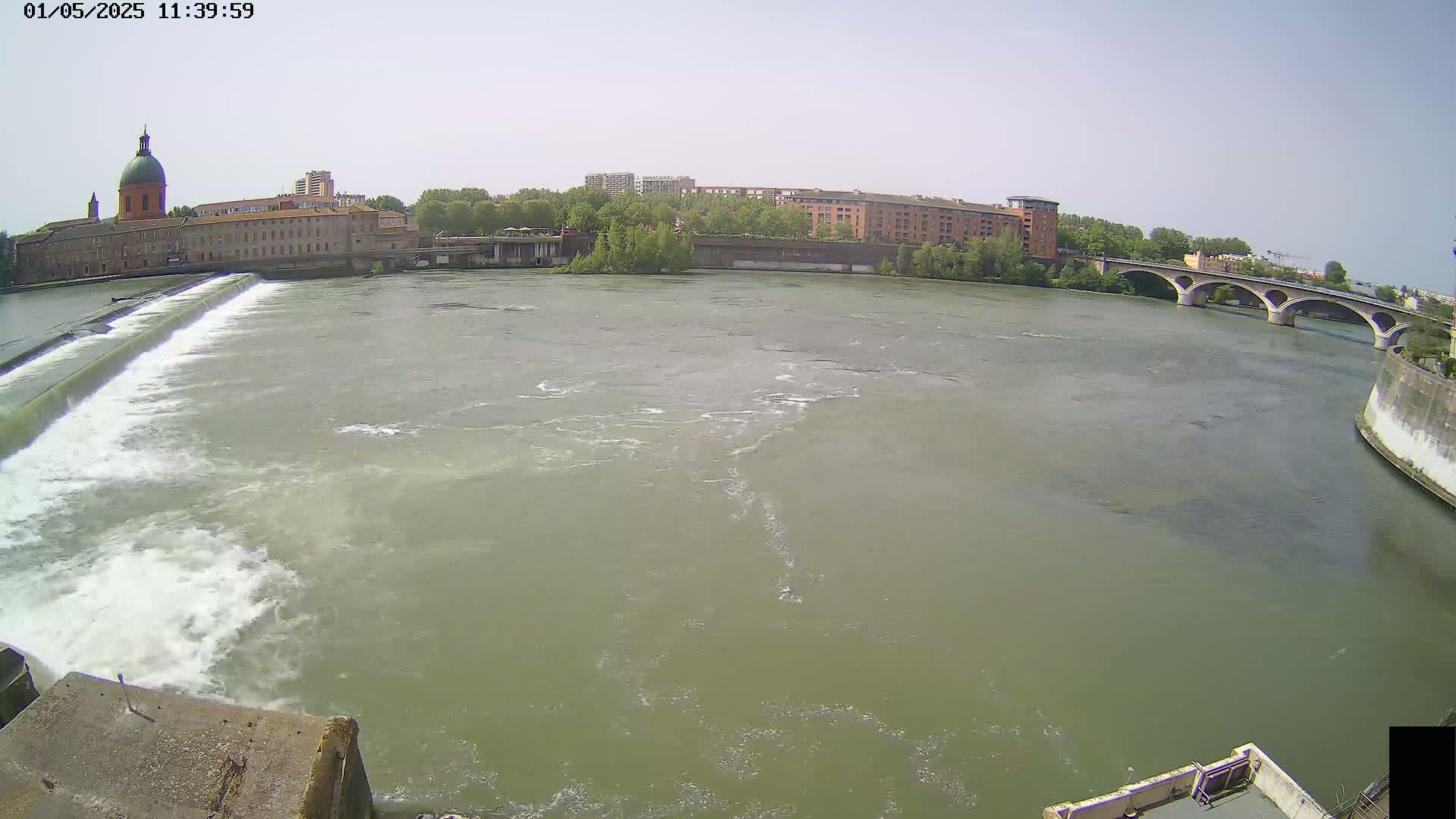 A wide river flows under a stone bridge on a sunny day, past a dam and buildings including a domed structure.