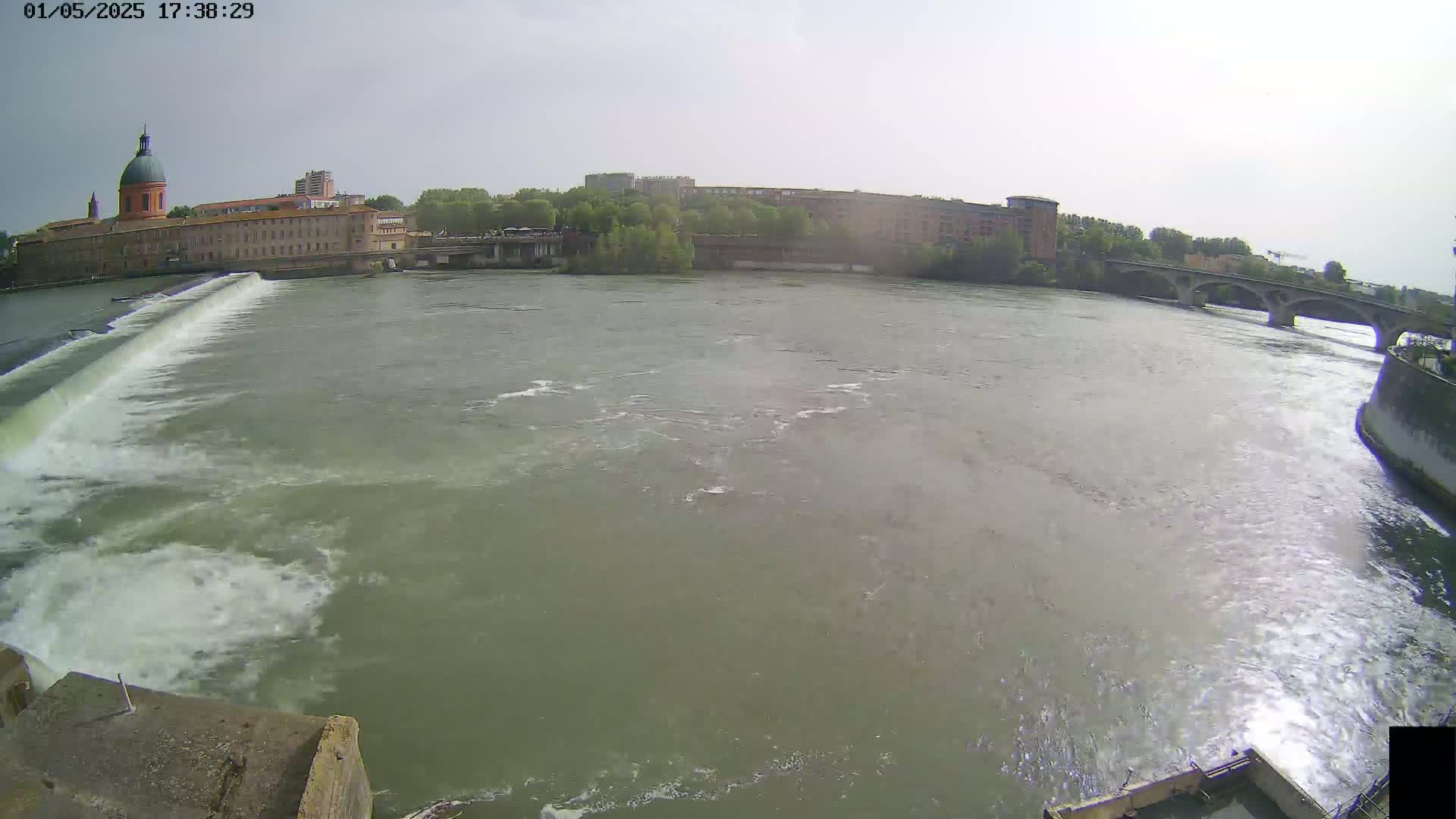 A wide shot shows a river flowing past a dam and under a bridge, with buildings and trees visible on the far bank under a partly cloudy sky.