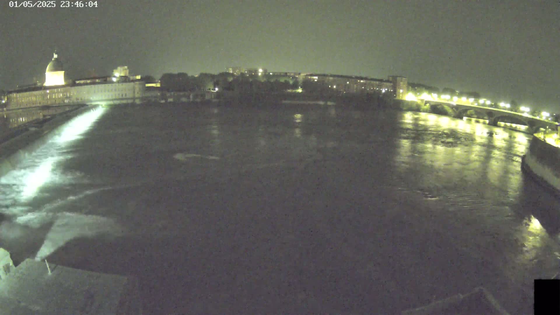 A wide nighttime shot shows a river flowing past a building with a dome and a bridge, with water flowing over a dam in the foreground.