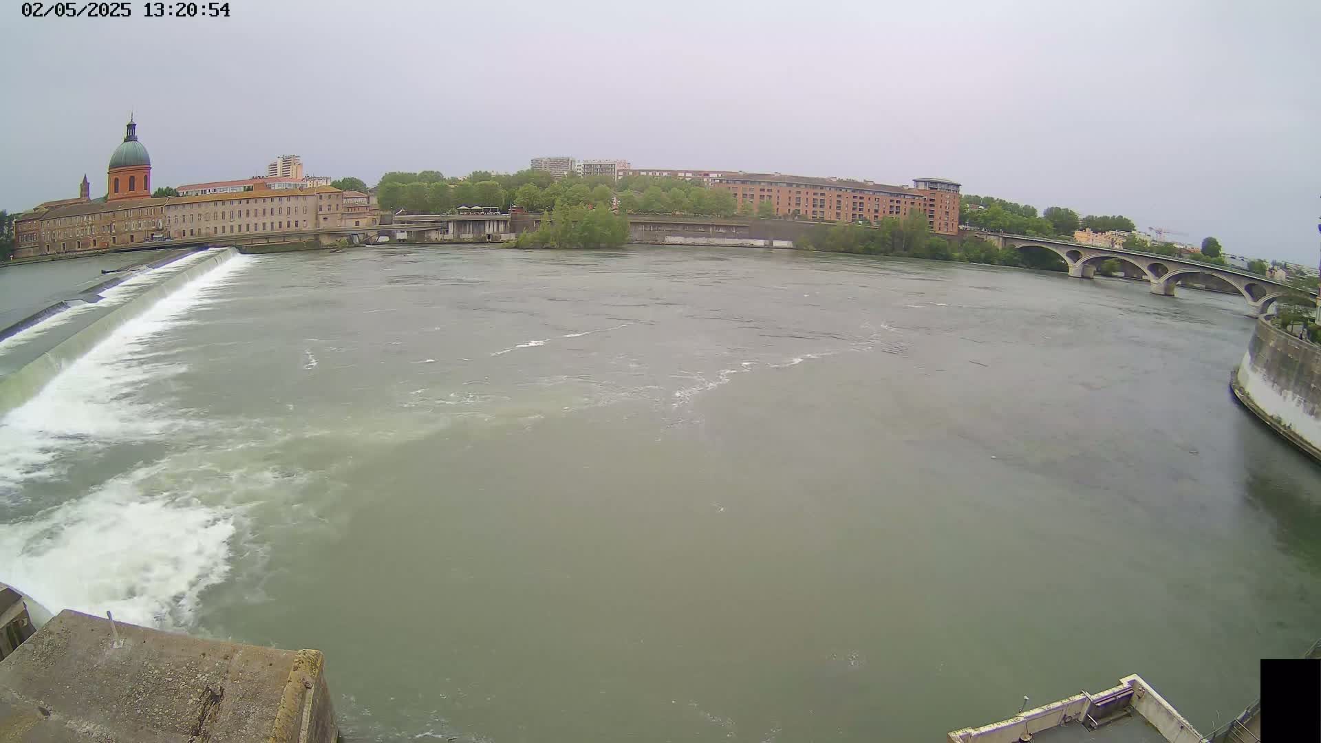A wide, murky river flows past a dam and under a bridge, with buildings and trees visible on the banks under an overcast sky.