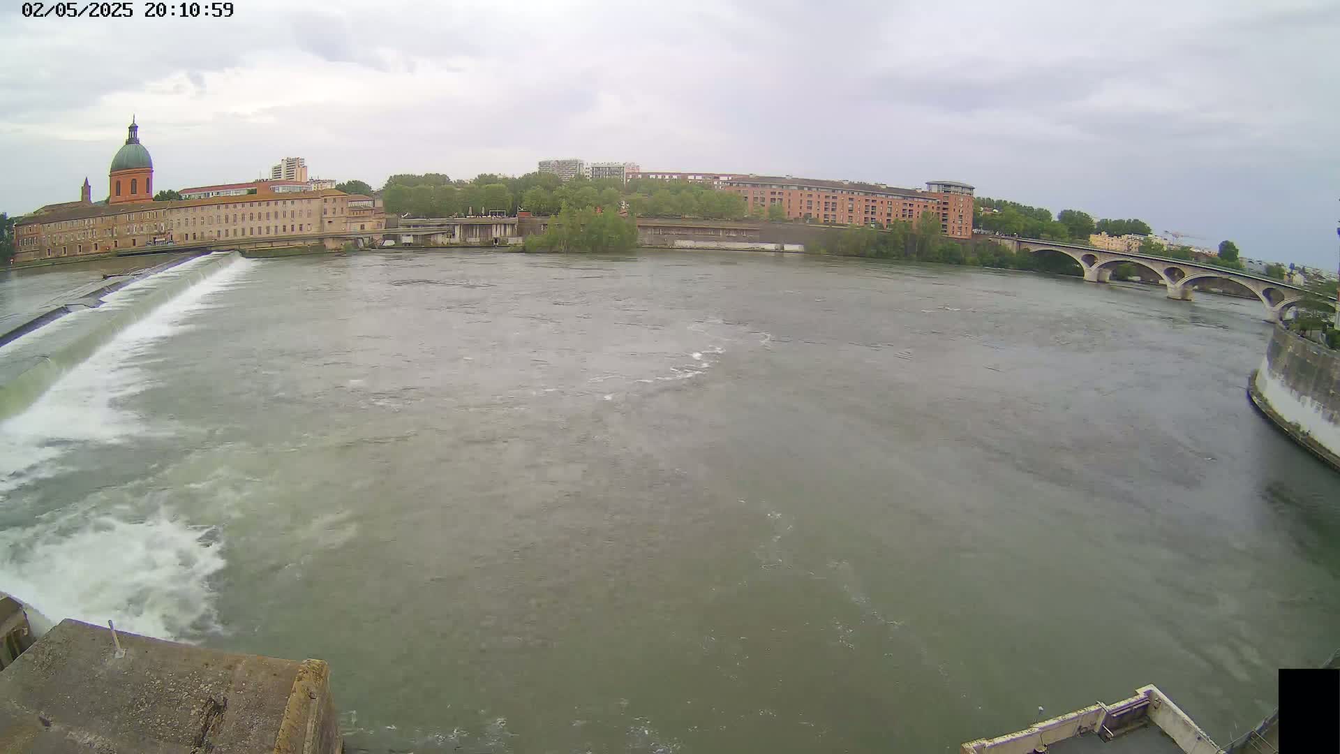 A wide shot of a river flowing past a dam and under a bridge on an overcast day, with buildings visible on the far bank.