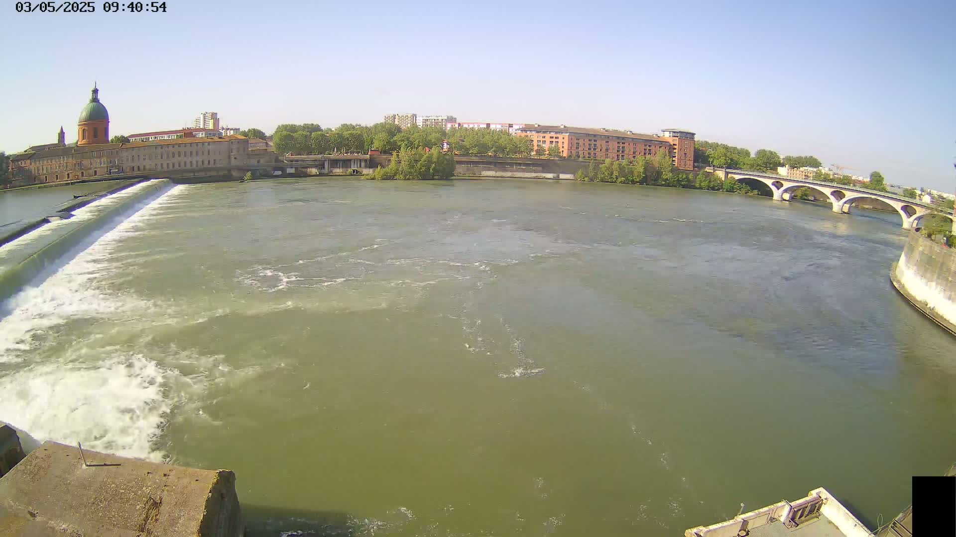 A wide shot on a sunny day shows a river flowing past a dam, with buildings and a bridge visible in the background.