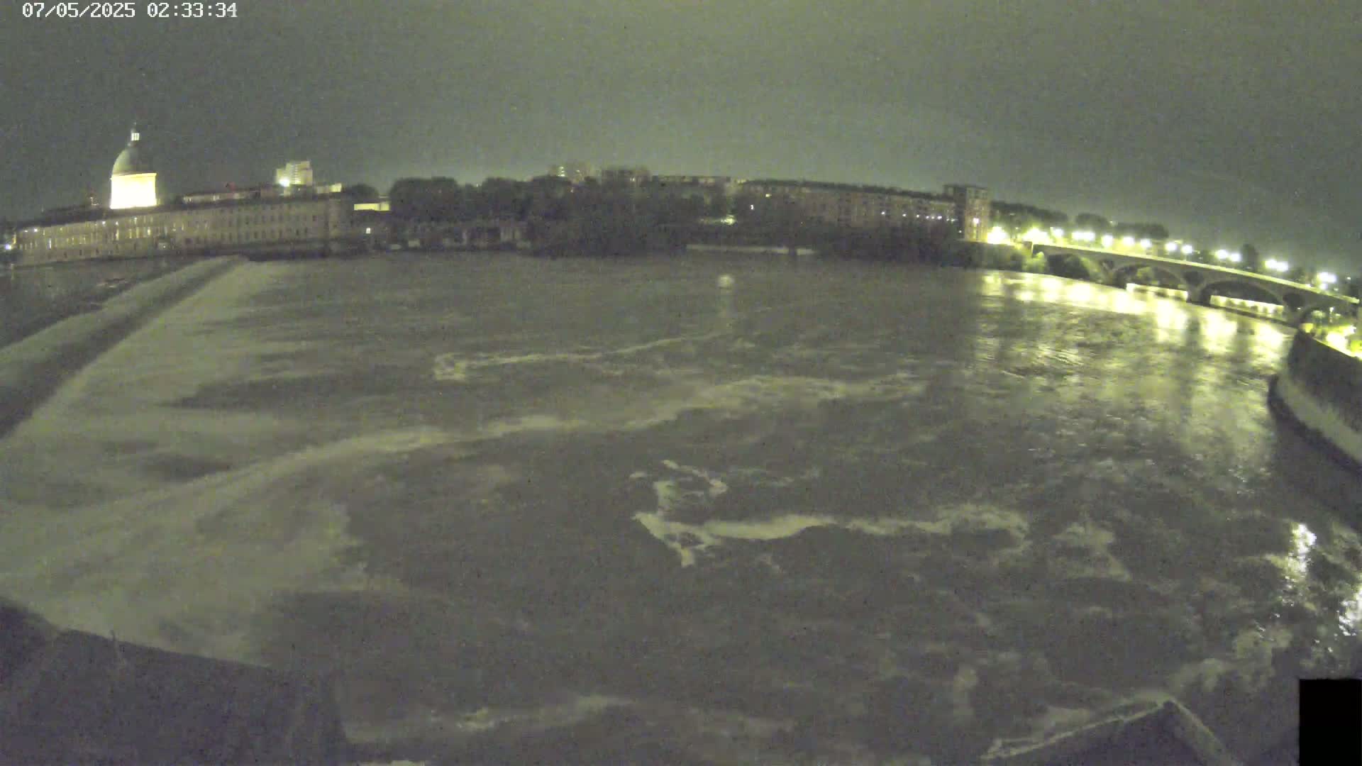 A nighttime view of a river flowing past city buildings and a bridge under a dark sky.