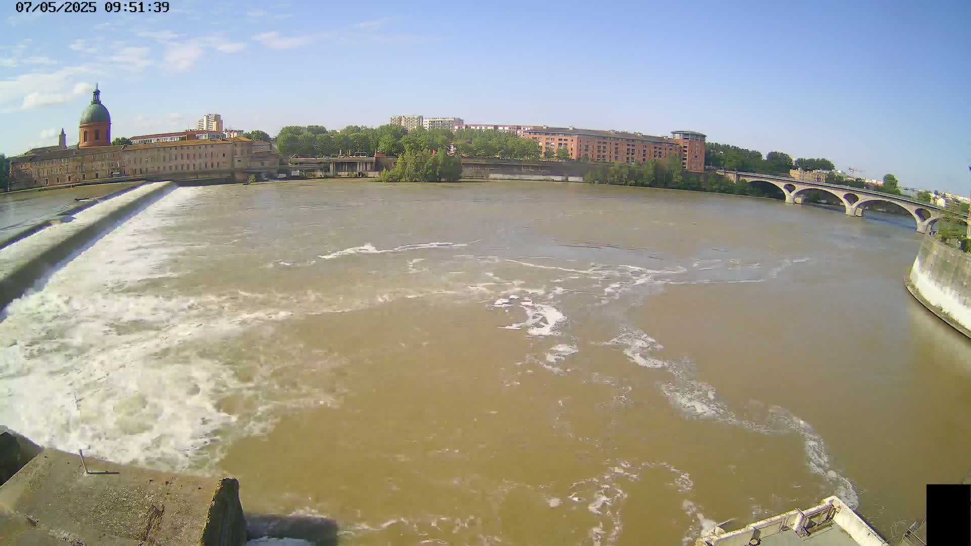 A wide shot of a muddy river flowing over a dam, past buildings and a bridge under a clear blue sky.
