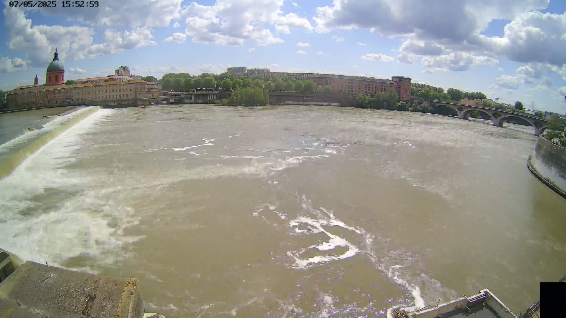 A wide shot of a river flowing rapidly over a dam, with a city and buildings visible in the background under a partly cloudy sky.