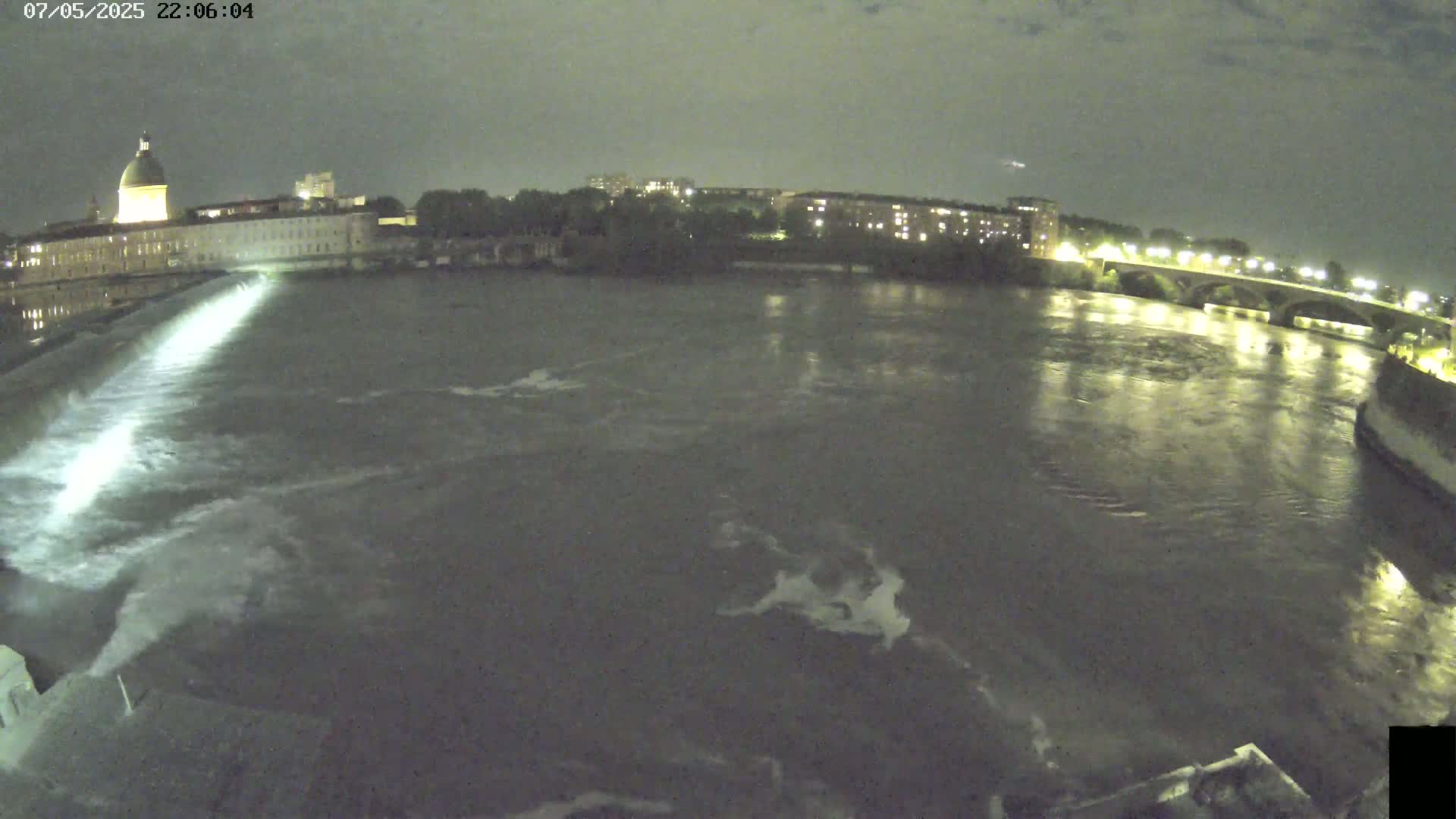 A nighttime view shows a river flowing past a building with a dome, under a dark, cloudy sky, with a bridge visible in the distance.