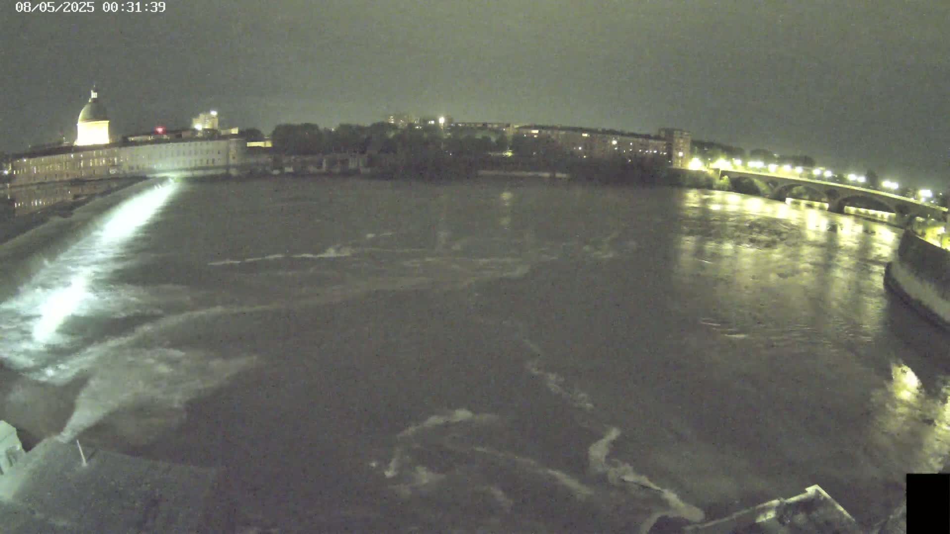 A nighttime view of a river flowing past a city with a bridge and a domed building, under a dark, cloudy sky.